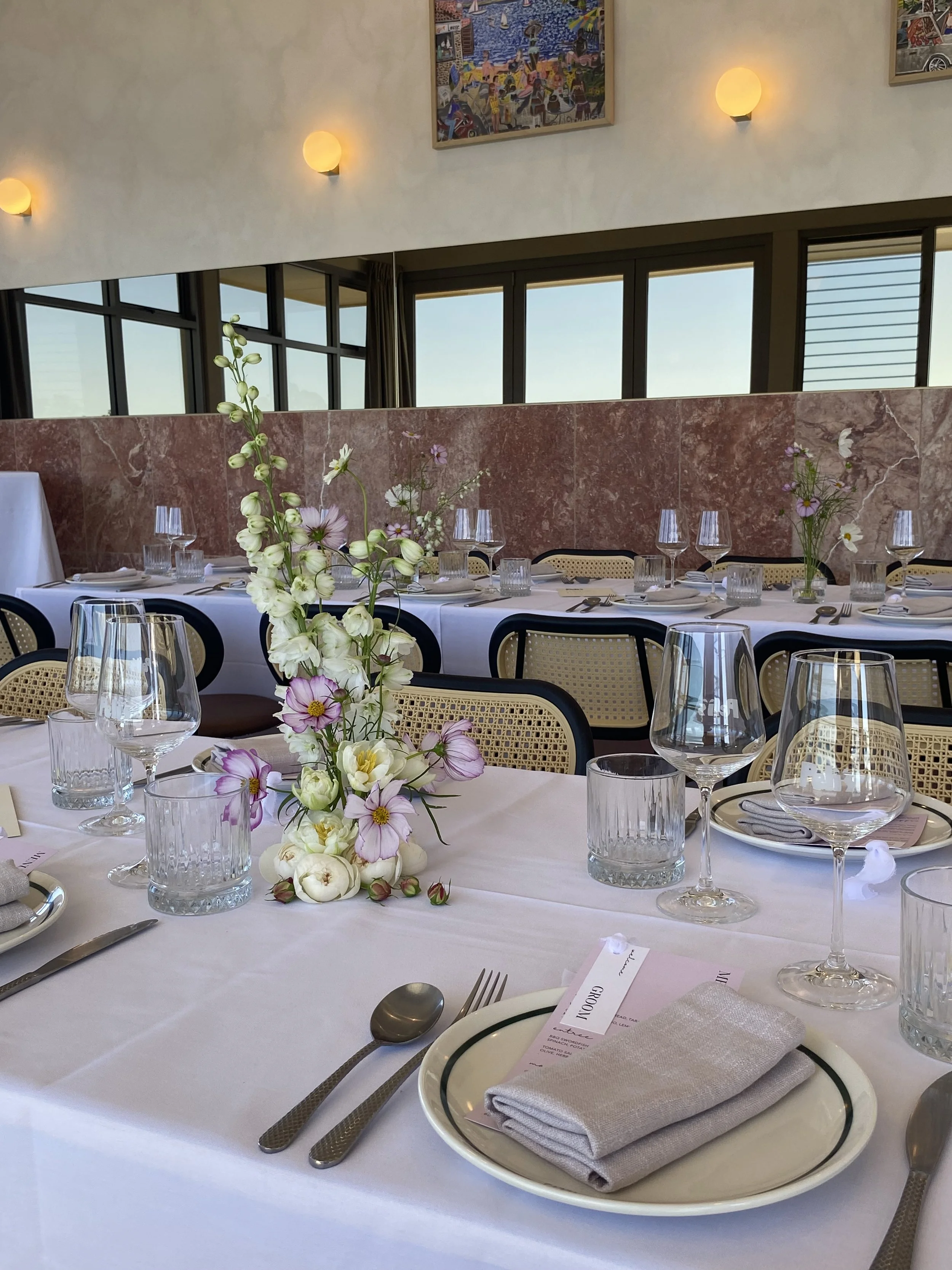Elegant dining table set with white tablecloth, featuring floral centerpieces, wine glasses, water glasses, cutlery, and neatly folded gray napkins on white plates with green rims in a well-lit restaurant with large windows and colorful artwork on th