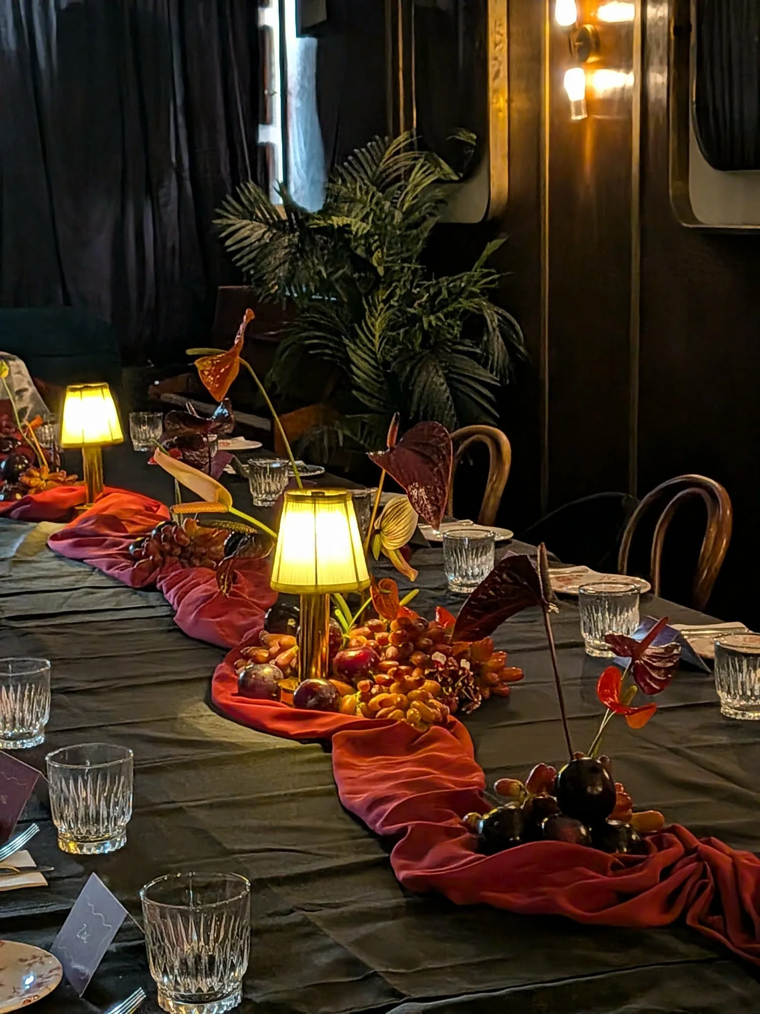 A dining table decorated with a red cloth, fruits, and flowers, with yellow lamps providing warm lighting in a dimly lit room with dark curtains and framed artwork on the wall.
