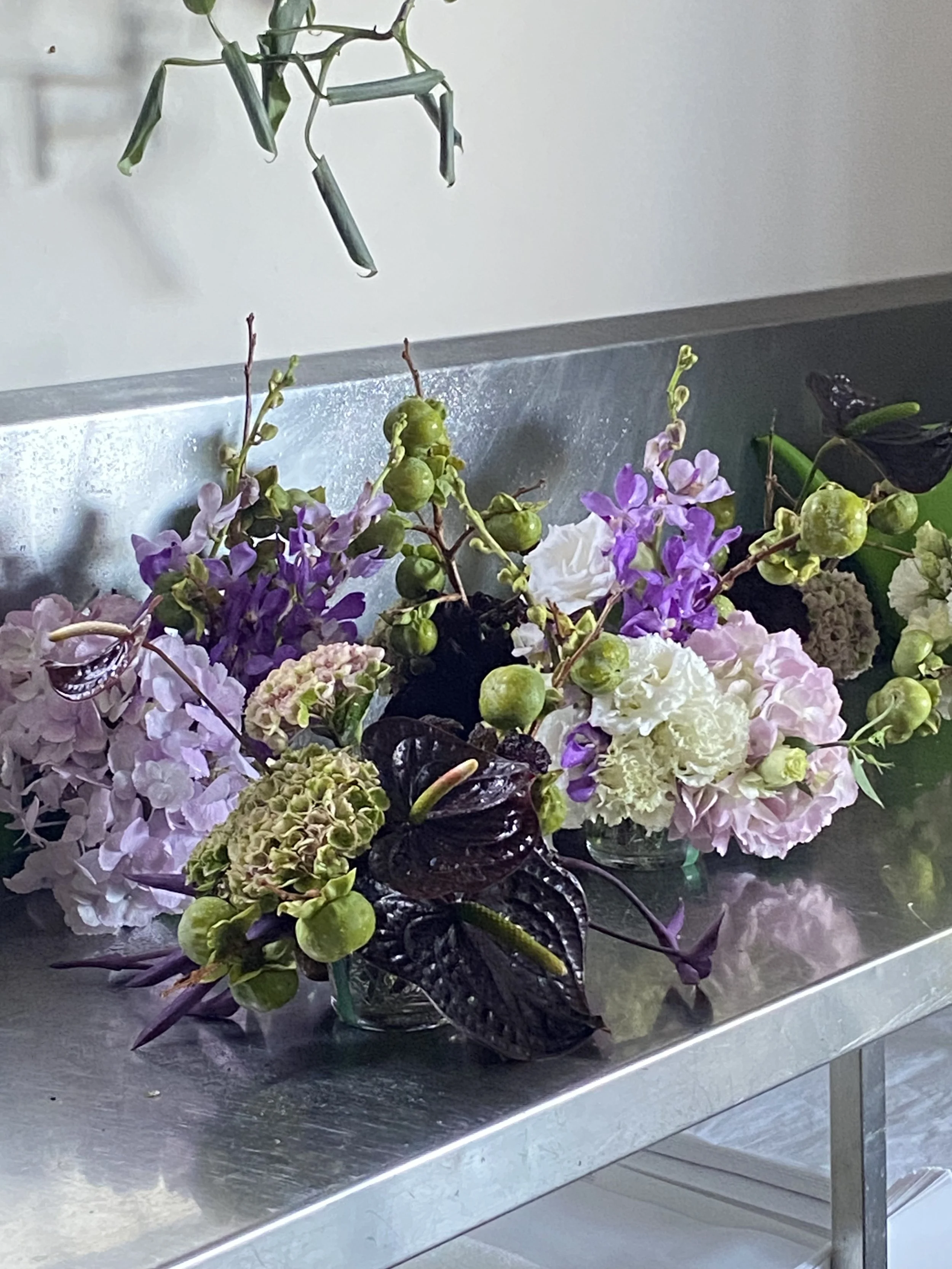 A metal table holding a transparent glass bowl filled with a colorful flower arrangement featuring hydrangeas, purple stock, green berries, and dark purple leaves, with a plant hanging above in a white room.