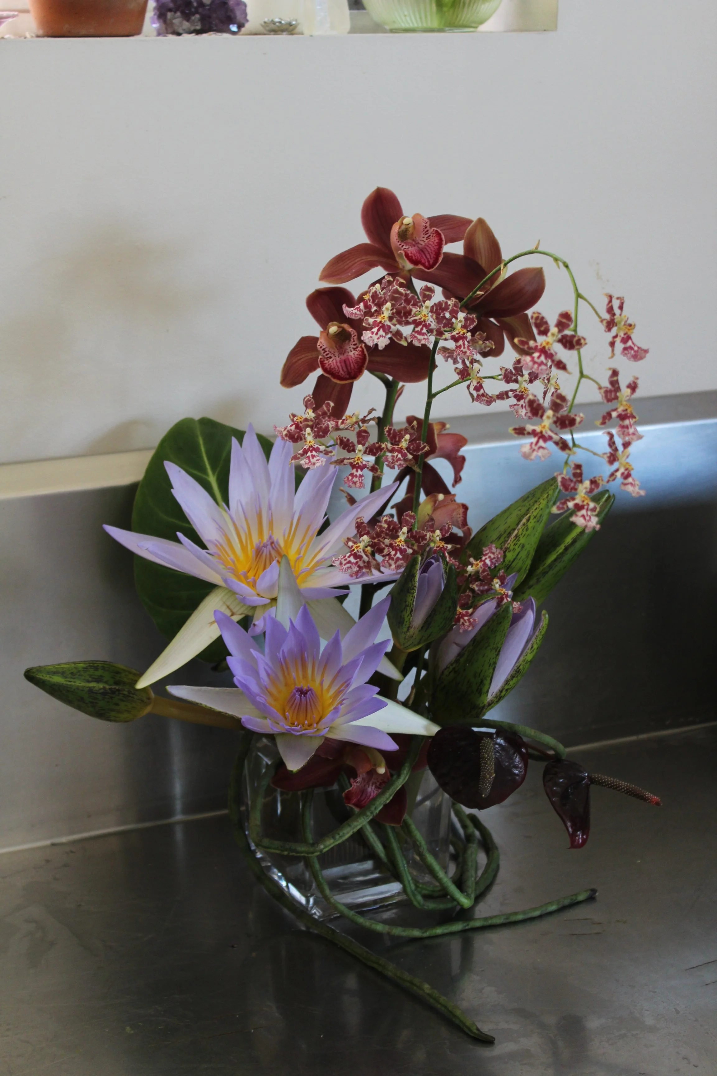 A floral arrangement featuring a purple water lily, red orchids, and other flowers in a glass vase on a metal surface with a white wall background.