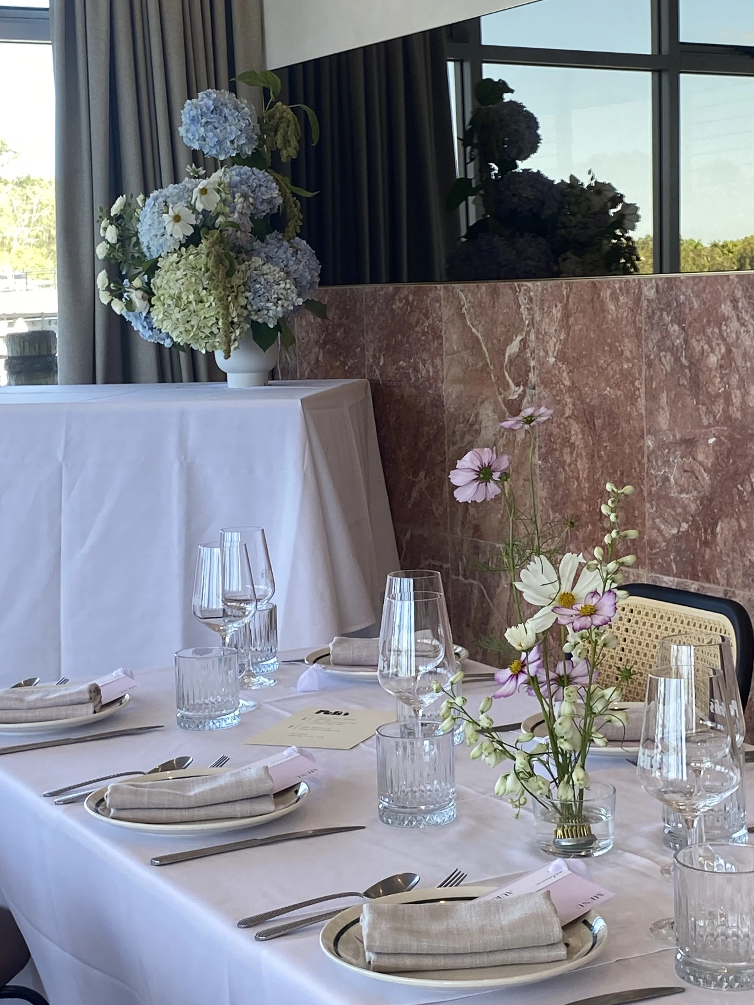 A formal dining table set with white tablecloth, plates, cutlery, napkins, water glasses, and a floral centerpiece with light-colored flowers, situated in a room with large windows and curtains.