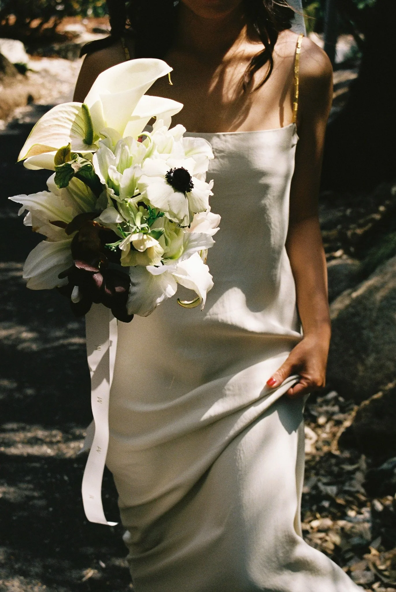 A woman in a white dress holding a bouquet of white flowers, standing outdoors with a rocky ground and trees in the background.