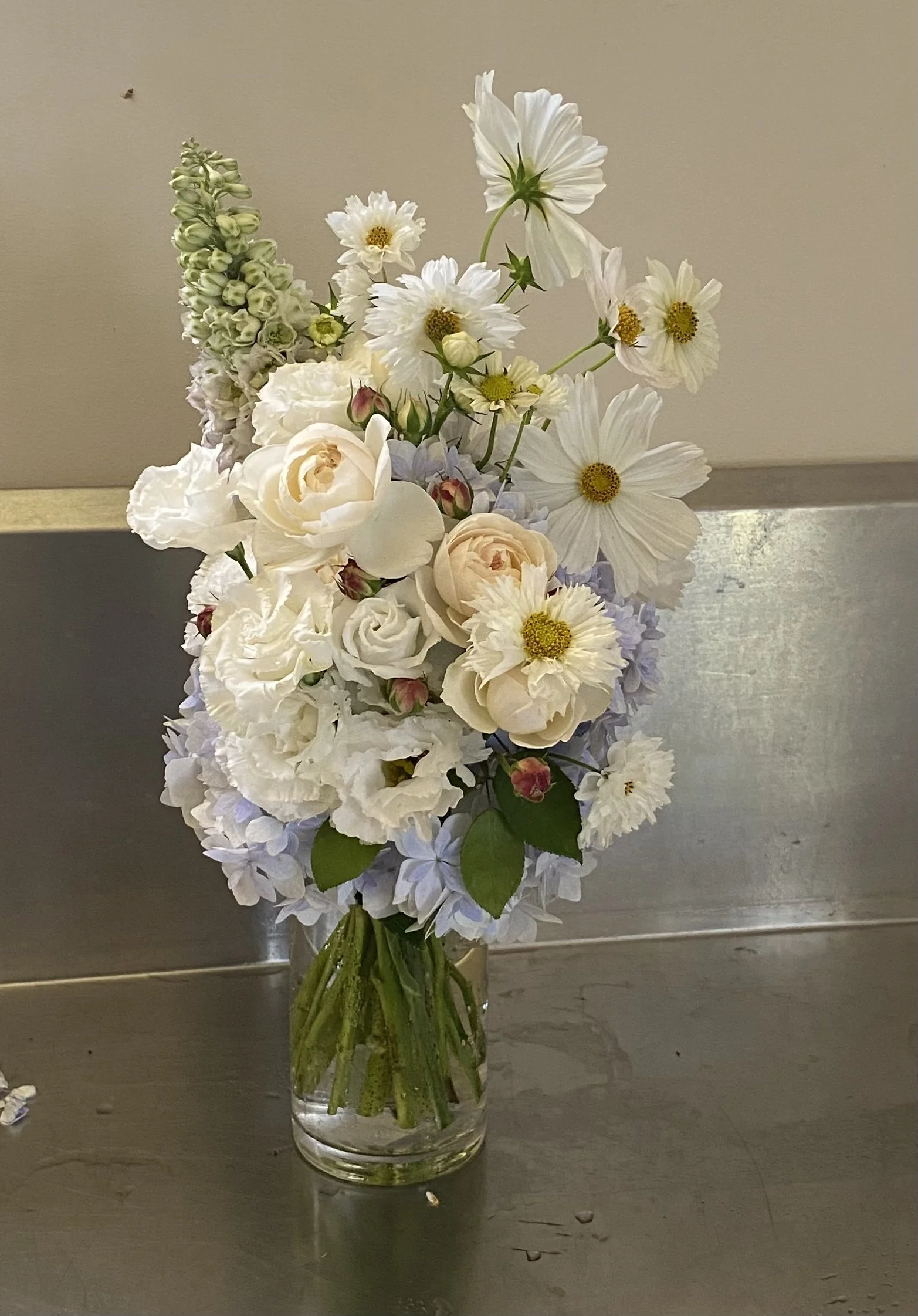 A clear glass vase with a bouquet of white flowers including roses, daisies, hydrangeas, and snapdragons, placed on a dark wooden surface against a plain beige wall.