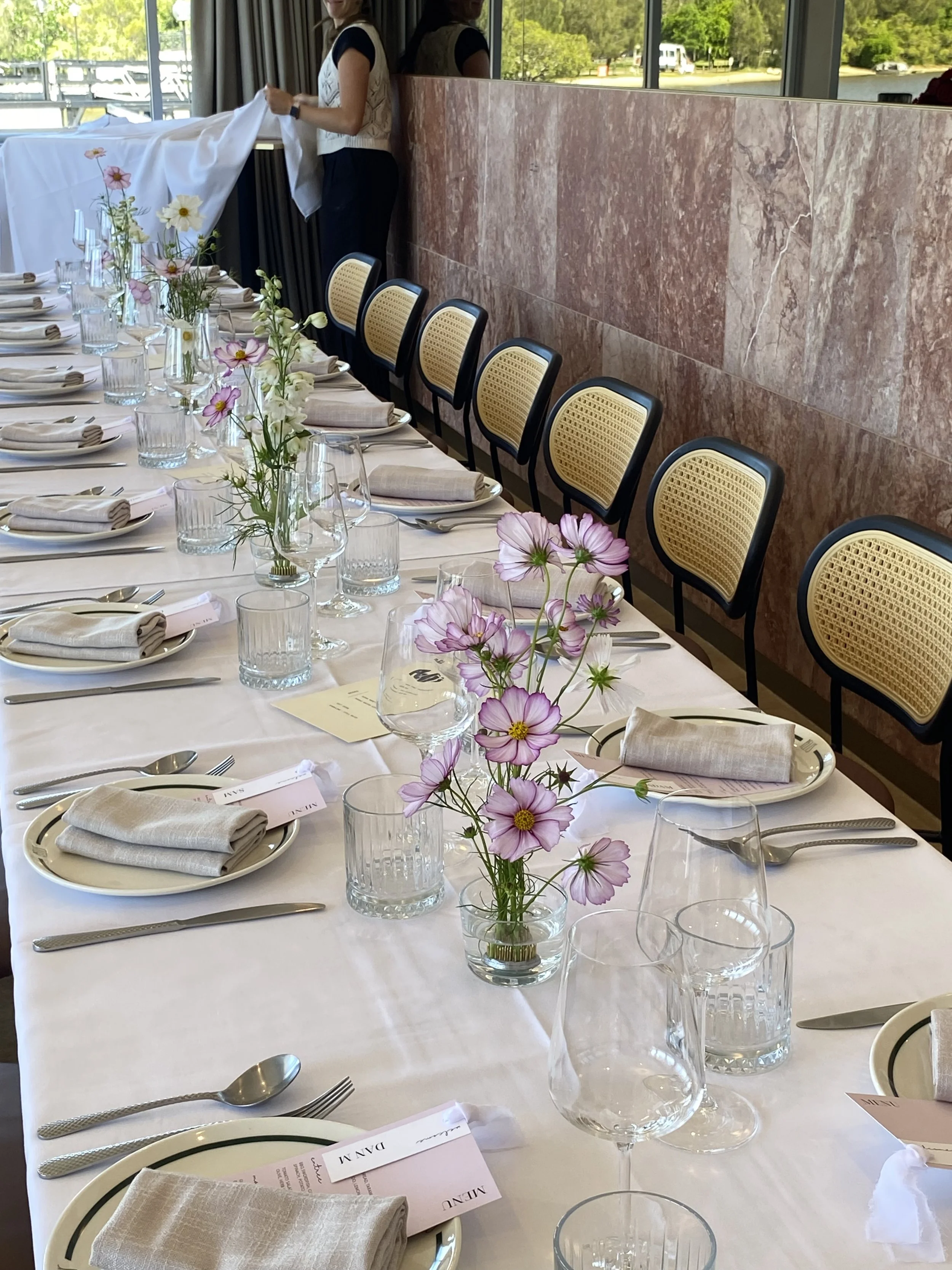 Elegant banquet table set with floral centerpieces, glassware, plates, silverware, and napkins at a formal event venue with large windows and outdoor scenery.