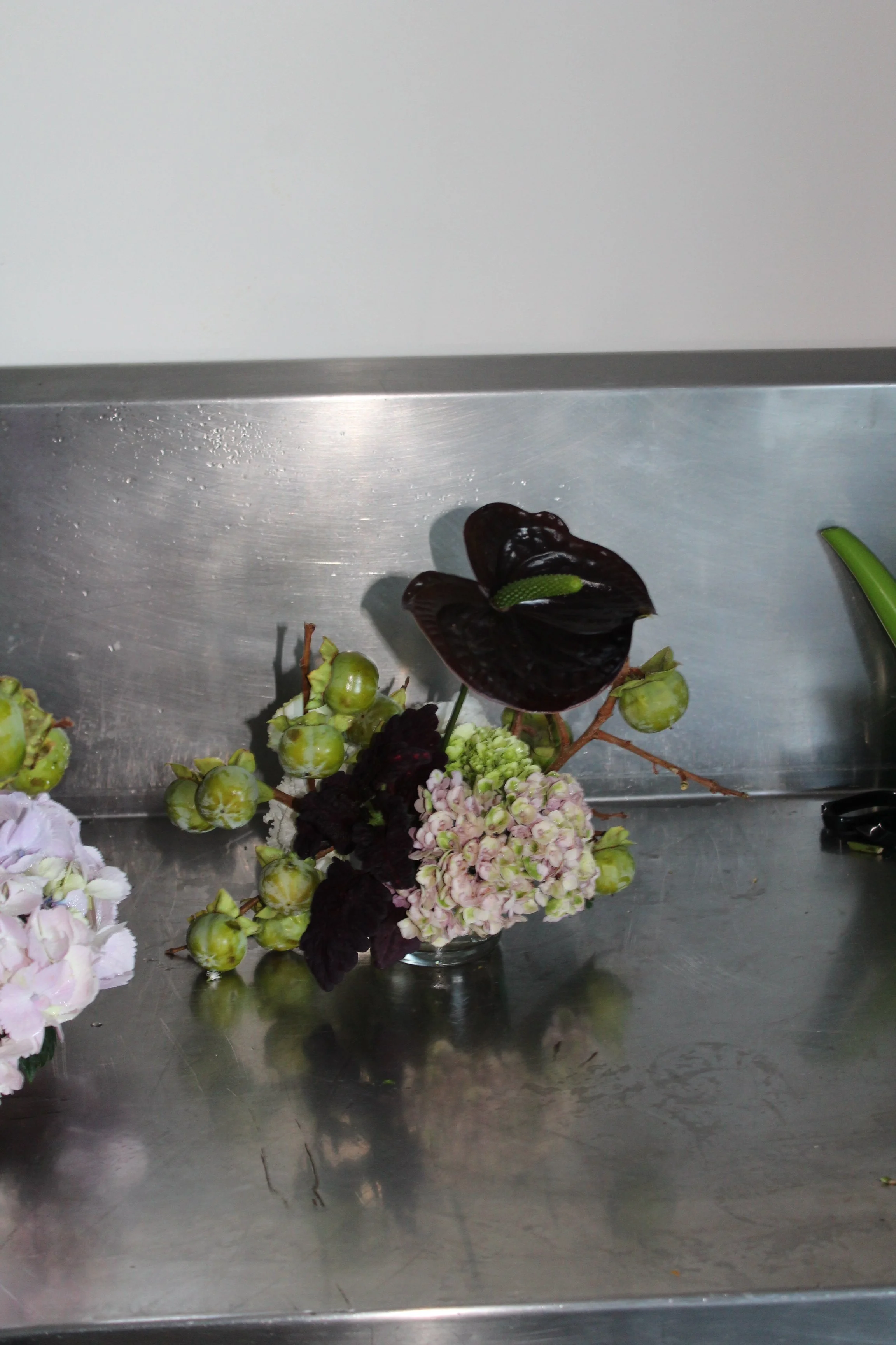 Flowers with dark purple and pink petals, green berries, and some greenery arranged on a metallic surface.