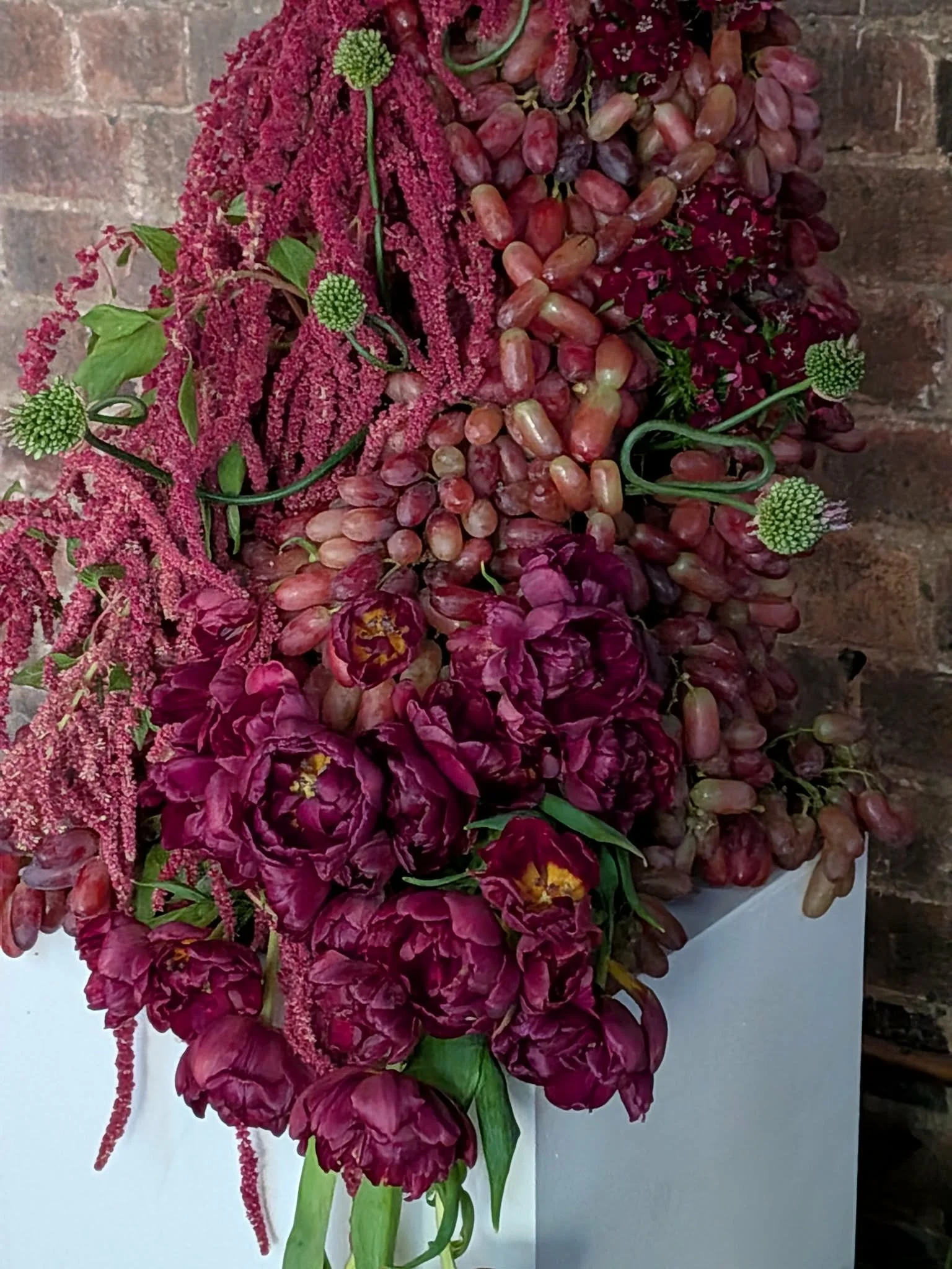 A floral arrangement featuring red flowers, green leaves, and small green spherical accents, set against a brick wall.