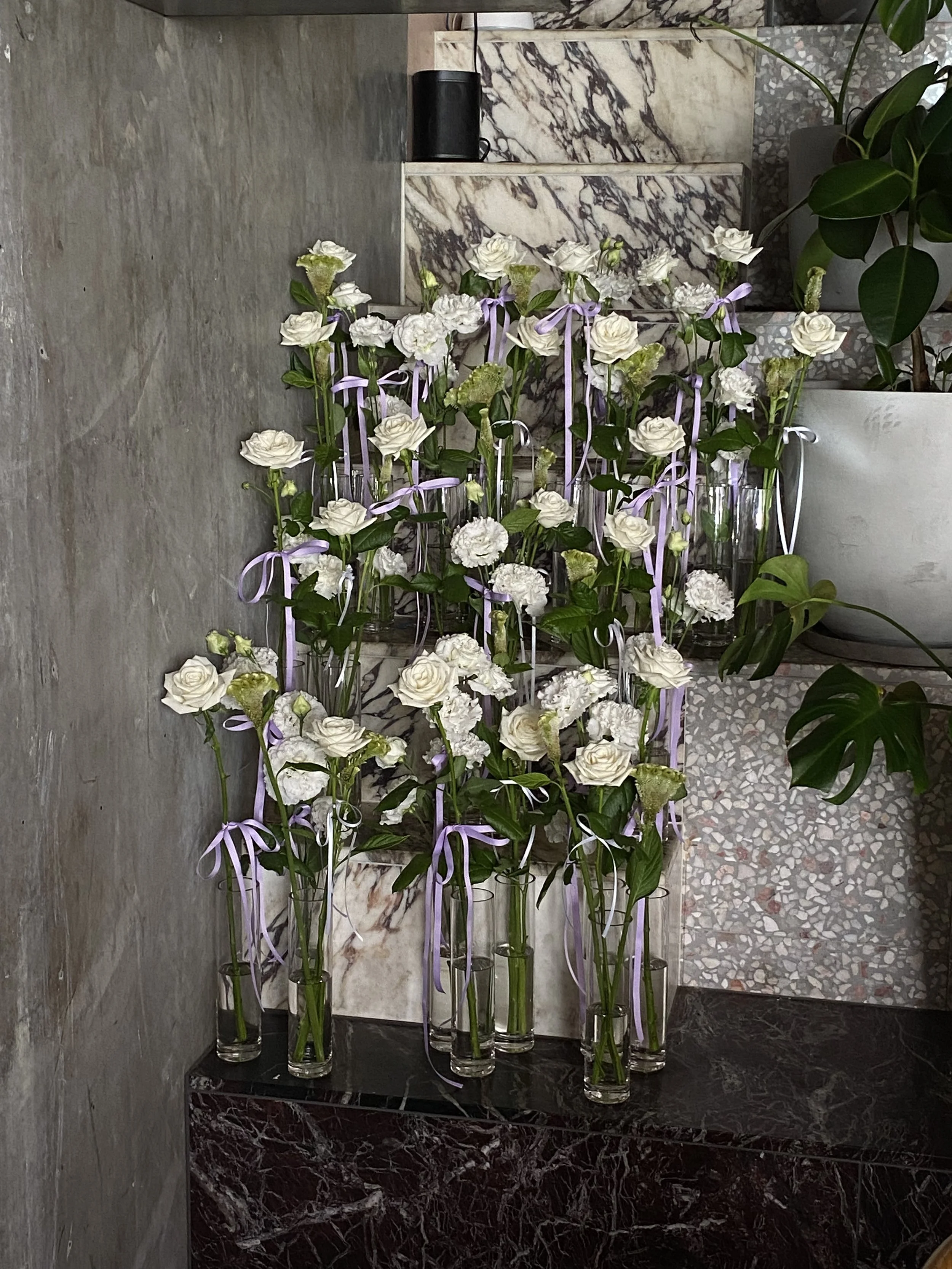 A display of tall glass vases filled with white roses and greenery, decorated with lavender ribbons, arranged on a dark marble surface against a marble wall with a potted plant nearby.