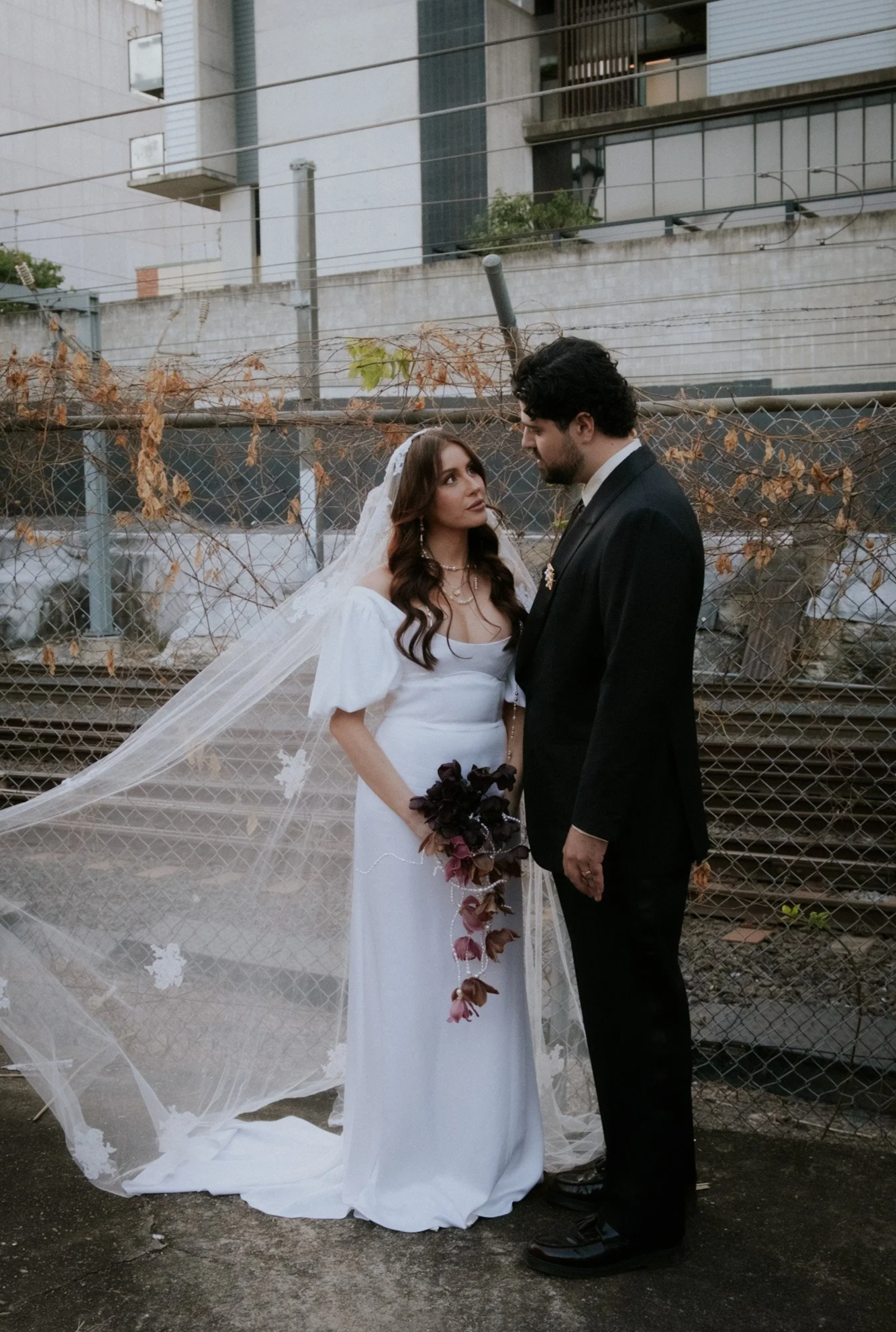 A bride and groom standing face-to-face outdoors, with a background of a chain-link fence covered in dried vines, and modern buildings behind it.