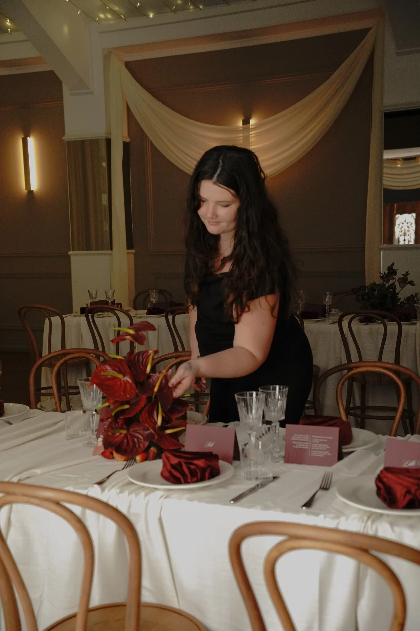 A woman with dark wavy hair in a black dress is setting the table at a formal event. The table has white tablecloth, multiple glasses, cutlery, pink napkins, and a floral centerpiece with red flowers. The background shows empty chairs, a wall with warm lighting, and elegant drapery.