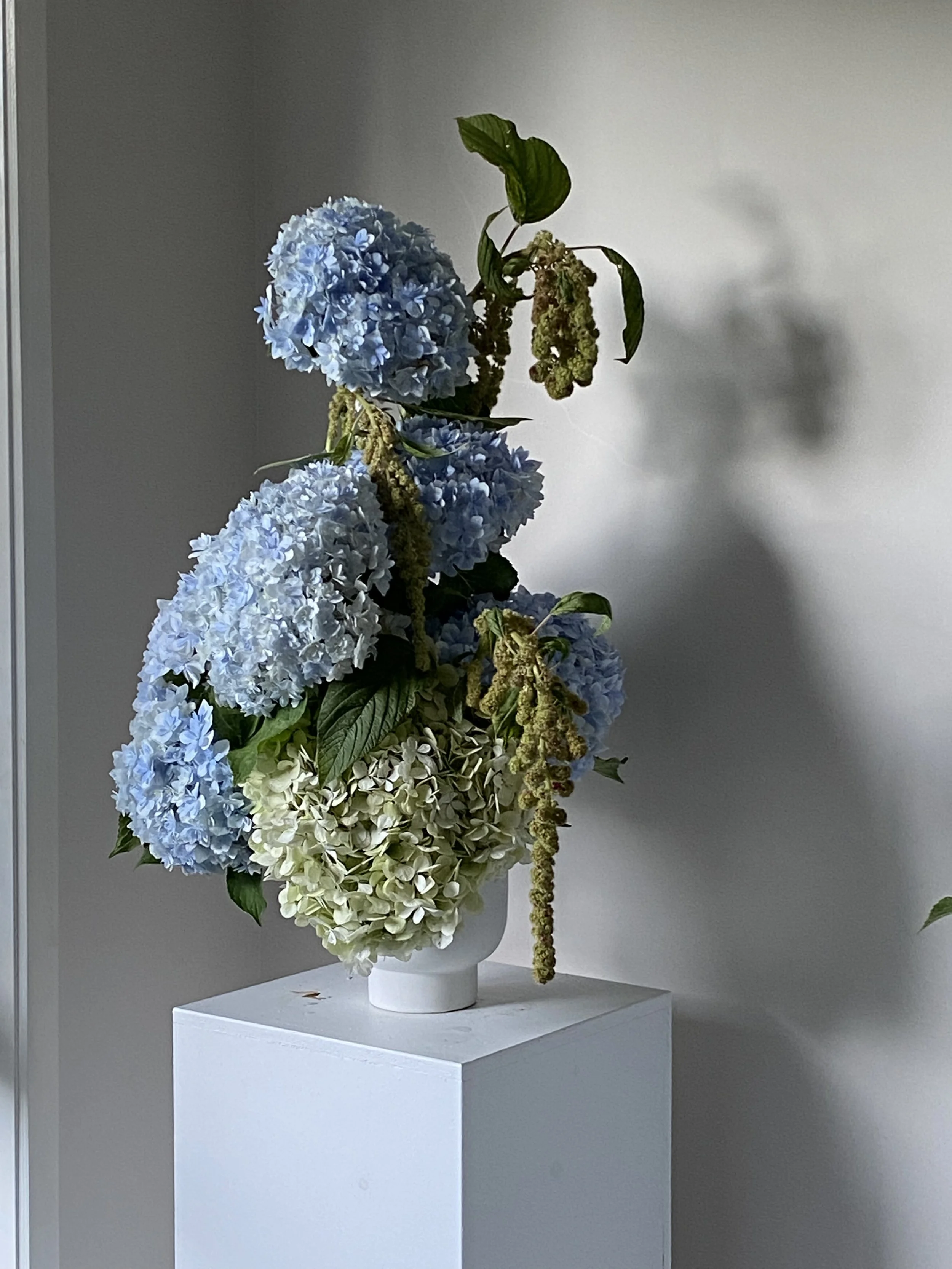 A tall white vase with white, blue, and cream hydrangea flowers on a white pedestal against a plain wall.