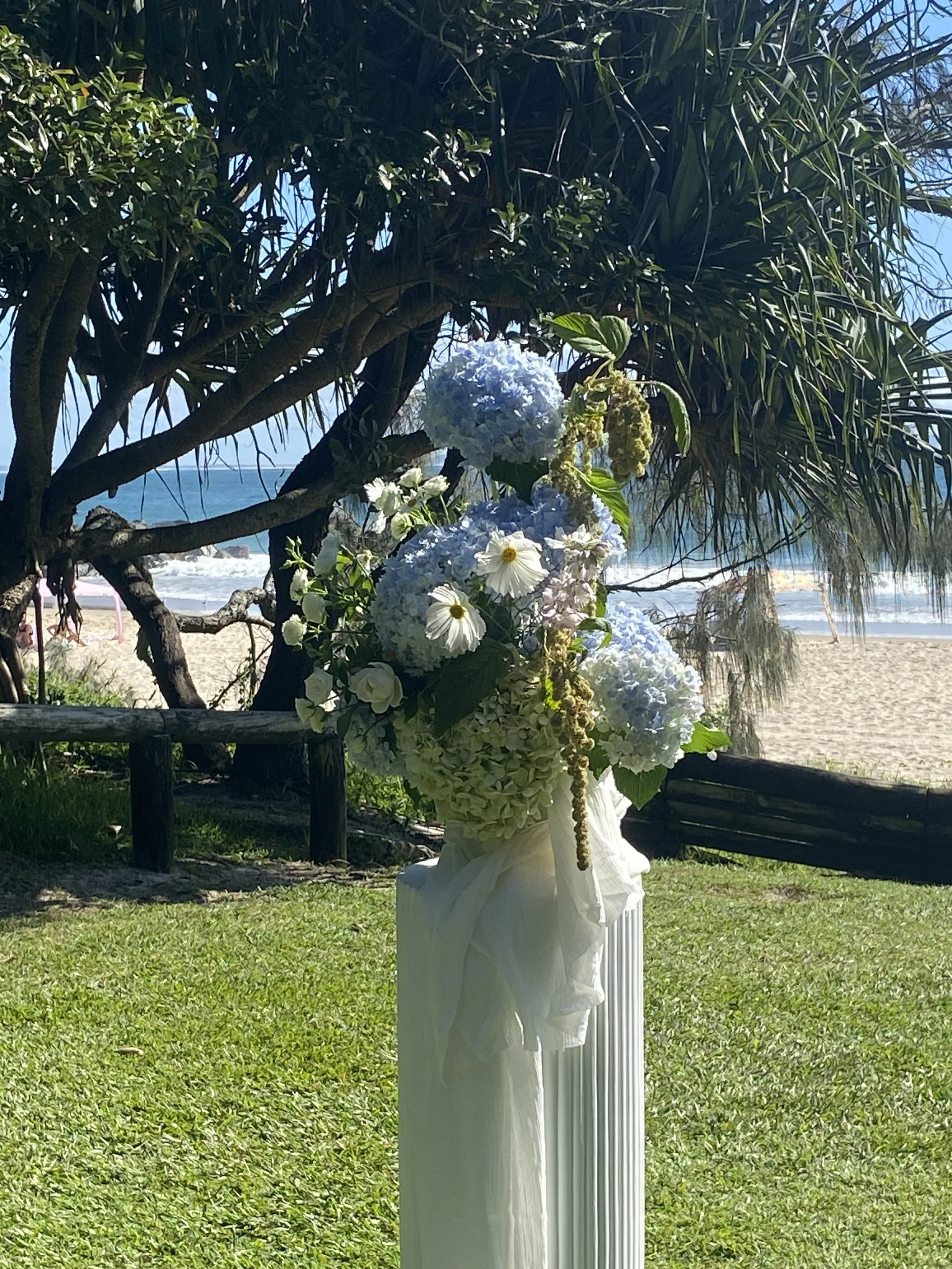 A decorated white pedestal with a floral arrangement of white and blue hydrangeas and white daisies, positioned on a grassy area near a beach with sand, rocks, and the ocean in the background, under a leafy tree.