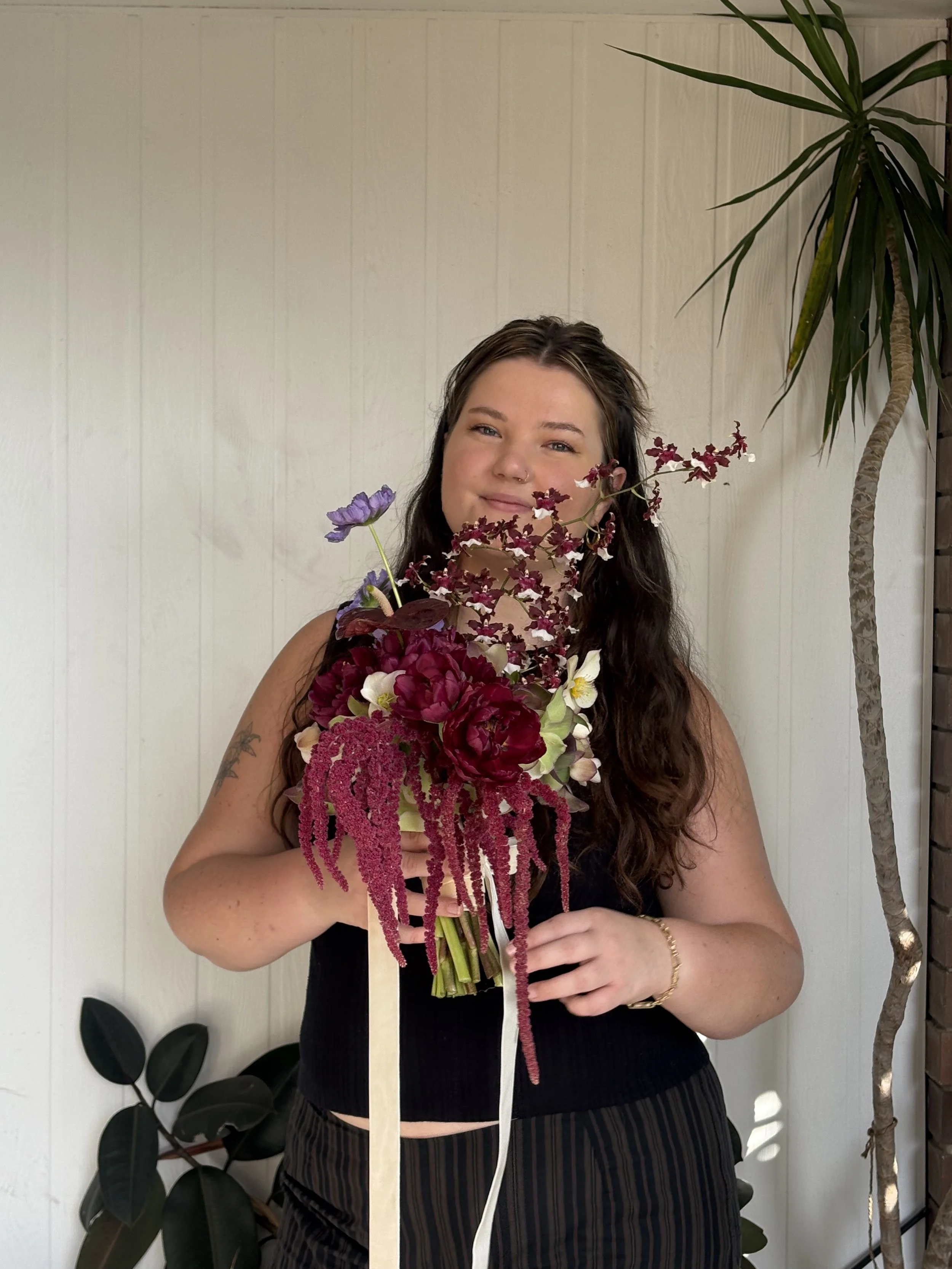 A woman with long dark hair holding a bouquet of pink, purple, and white flowers, standing in front of a white wall with plants to the side.