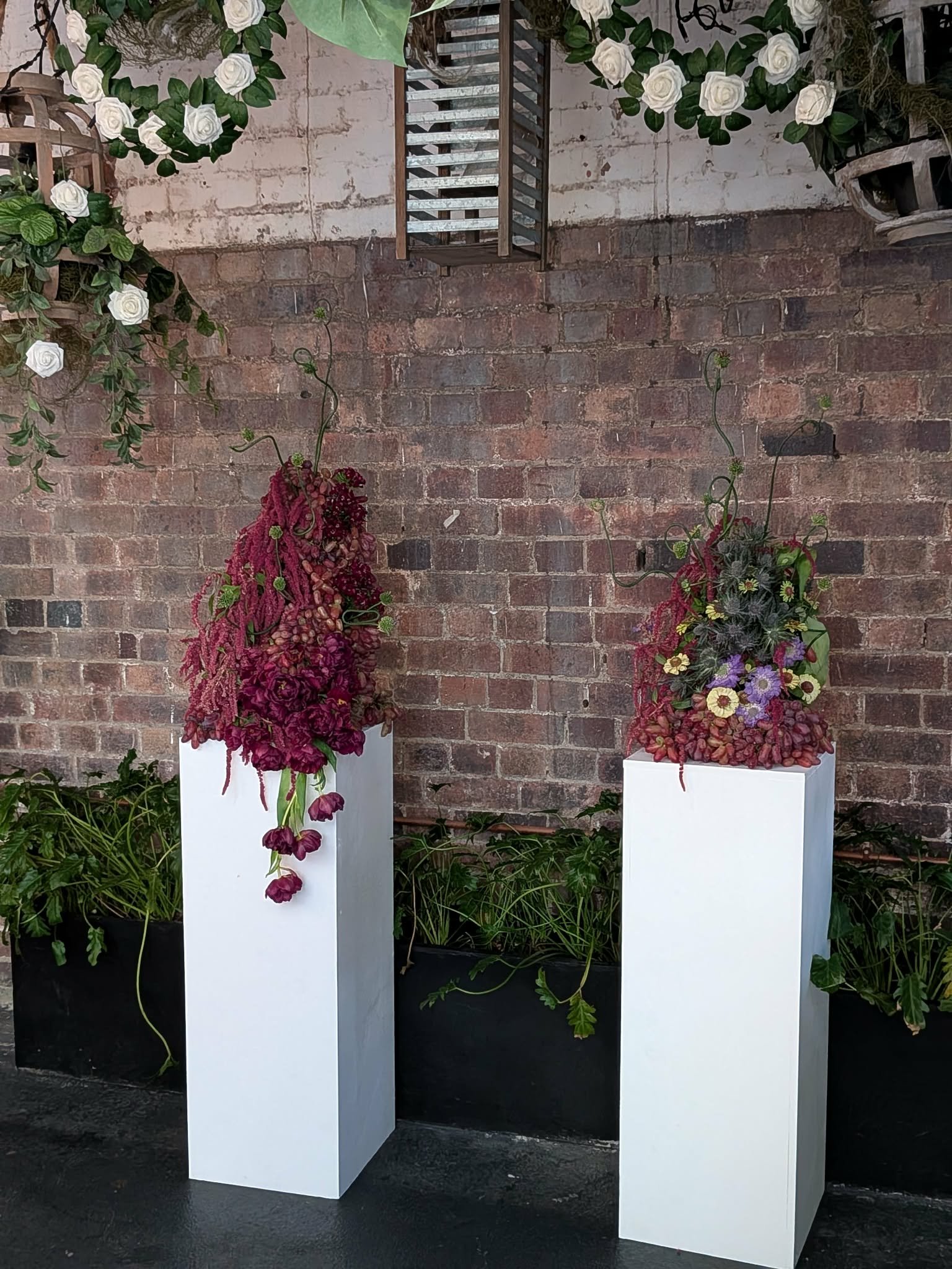 Three tall white pedestals with floral arrangements against a brick wall. Each arrangement features colorful flowers and greenery, with the left one having predominantly pink and purple flowers, and the right one with a mix of yellow, purple, and pin