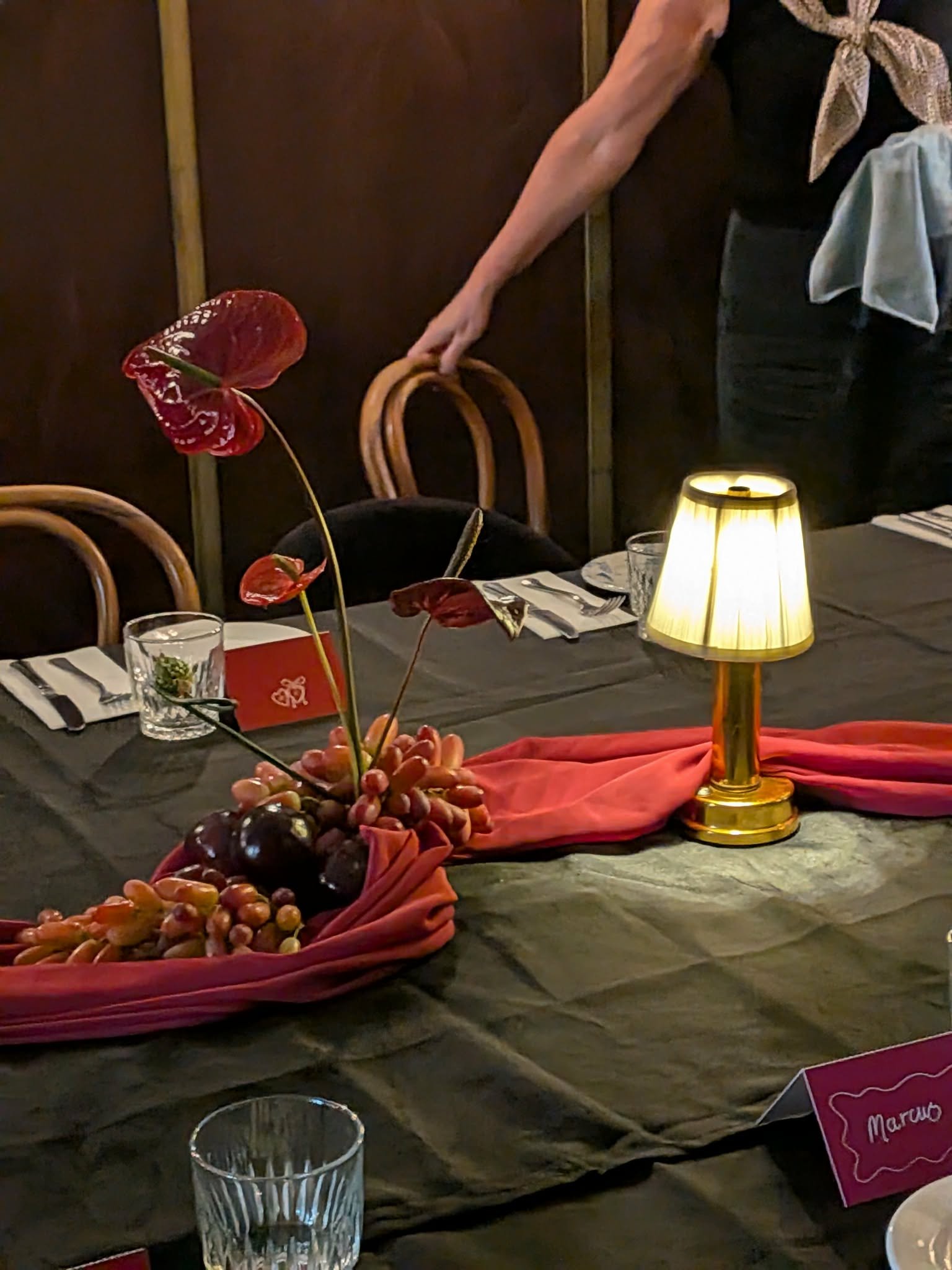 A dining table decorated for a celebration with a floral centerpiece of red anthurium flowers and berries, a small table lamp with a yellow lampshade, and a place setting with a menu card labeled 'Marcus'.