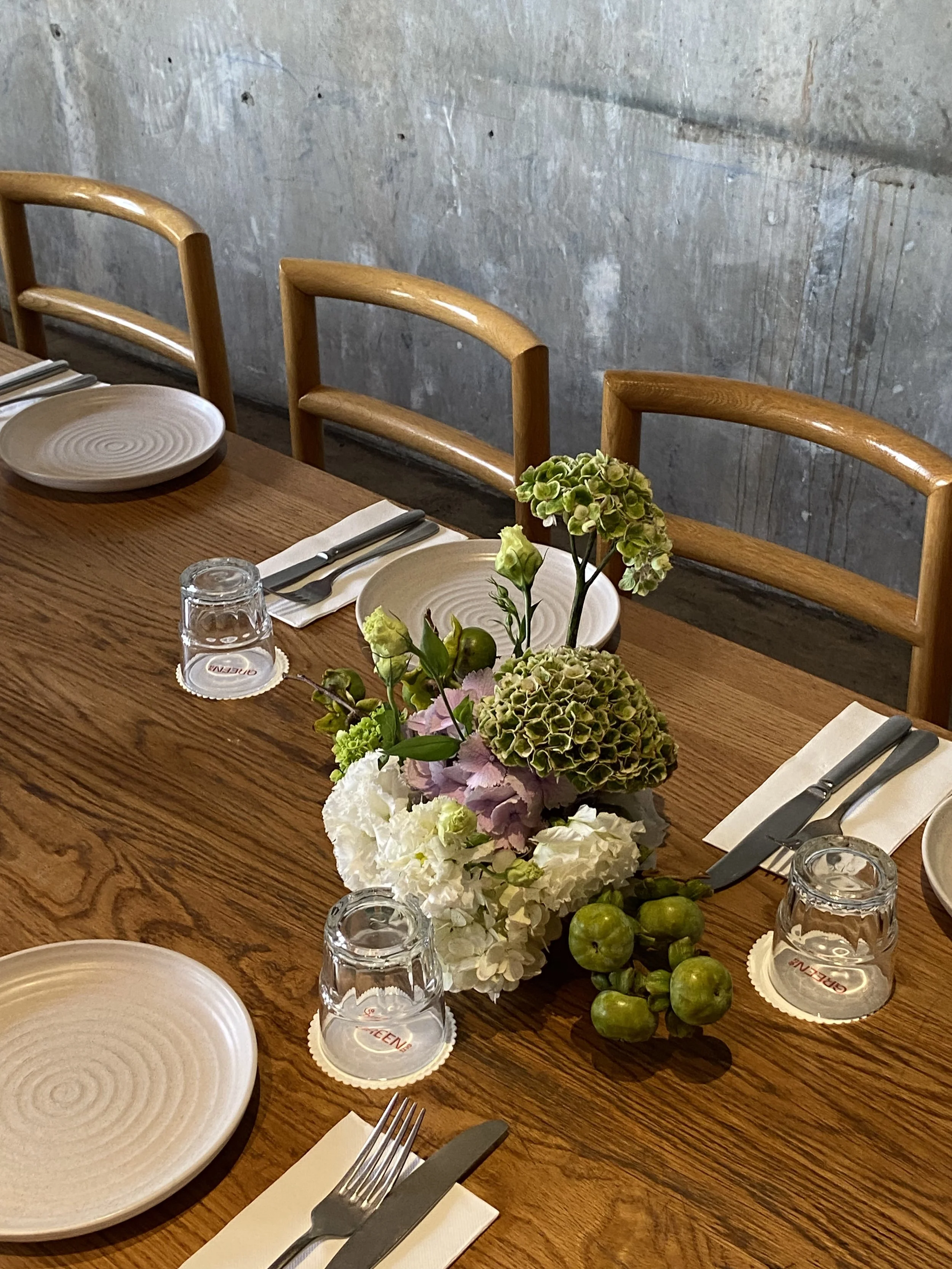 A wooden dining table set with white plates, silverware wrapped in white napkins, upside-down glass cups, and a floral centerpiece with hydrangeas, green apples, and other flowers, against a concrete wall background.