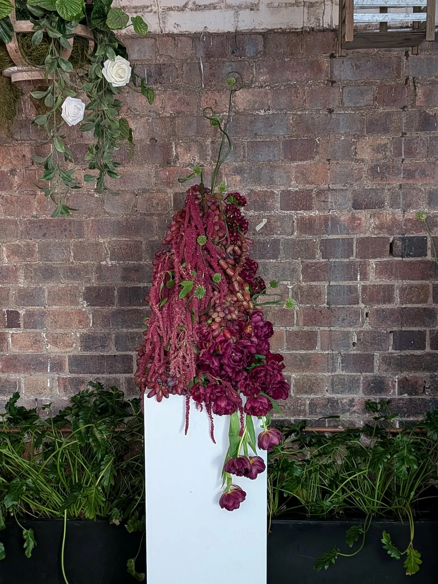 A floral arrangement with red and pink flowers and green accents, displayed on a white stand against an exposed brick wall, with greenery at the base.