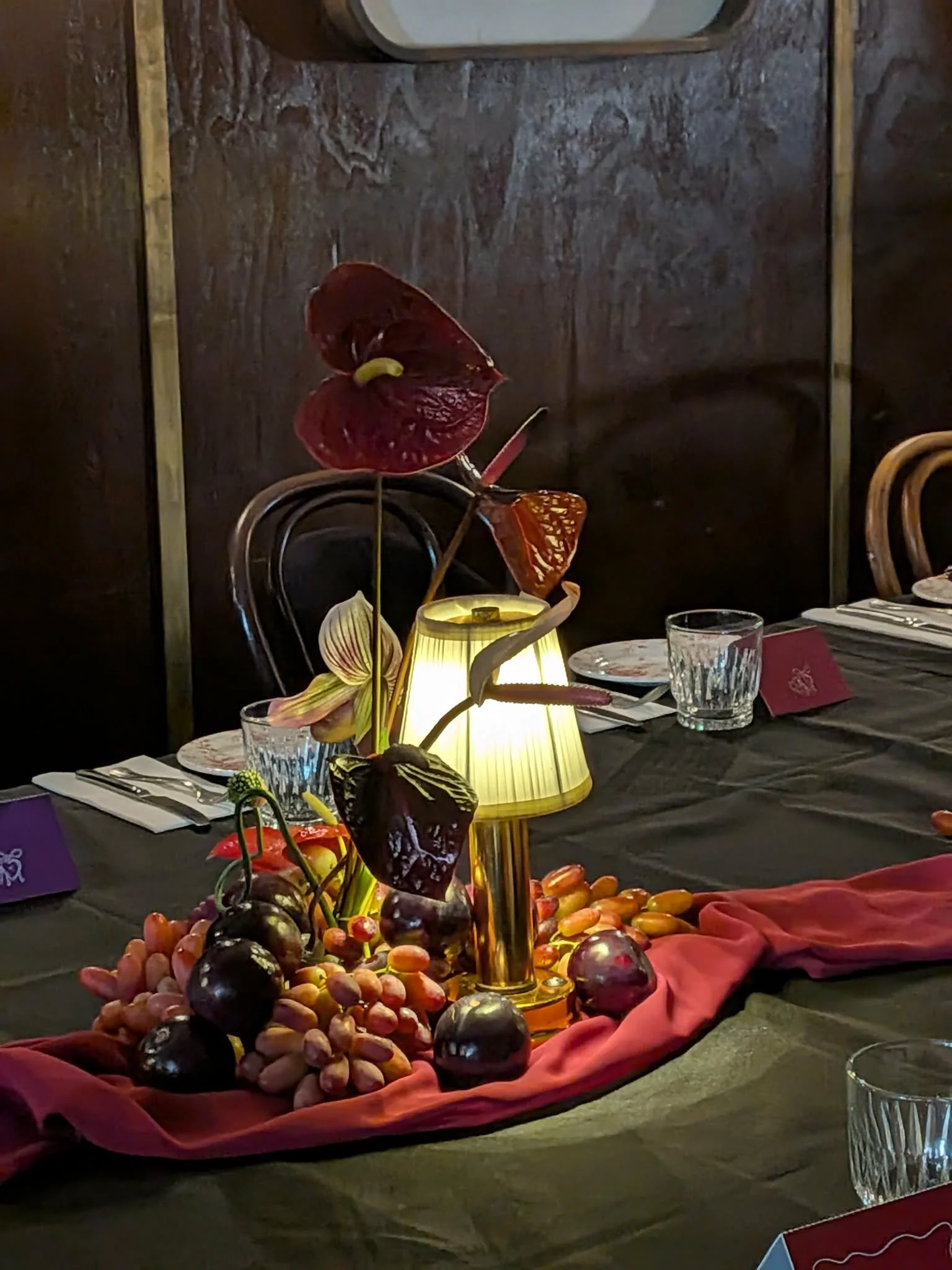 Decorative table centerpiece with assorted fruits, including grapes, on a cloth-covered table, illuminated by a table lamp in a restaurant setting.