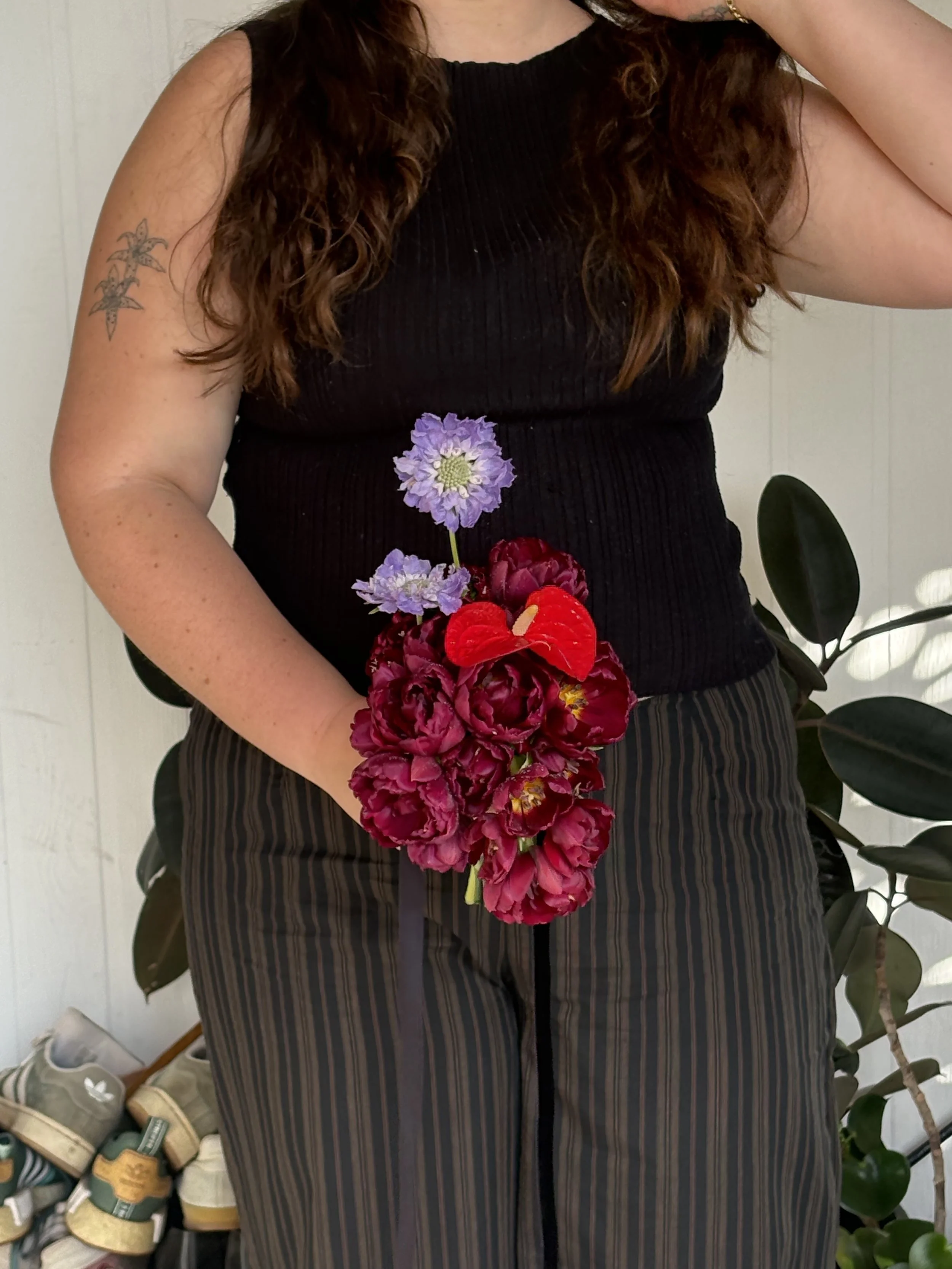 A woman with long, curly hair wearing a black sleeveless top and striped pants, holding a bouquet of pink and purple flowers, standing indoors with potted plants nearby.