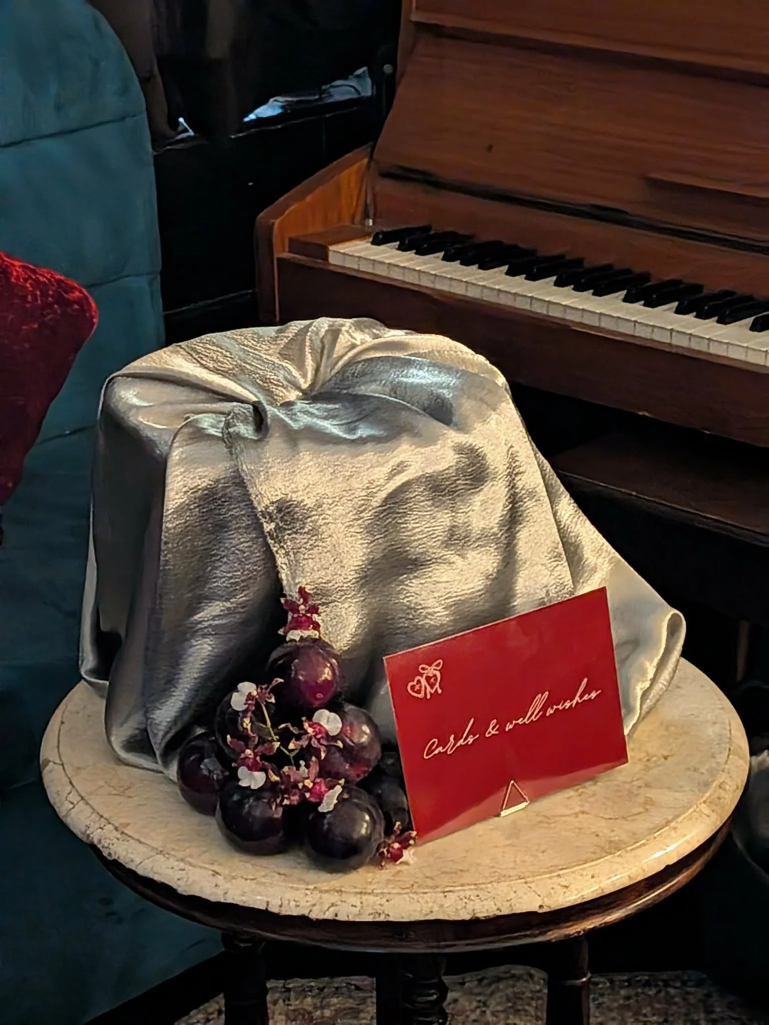 A marble table with a bunch of dark purple grapes, a sprig of red berries, and a card with handwritten text. In the background, an open upright piano and a blue velvet armchair are visible.