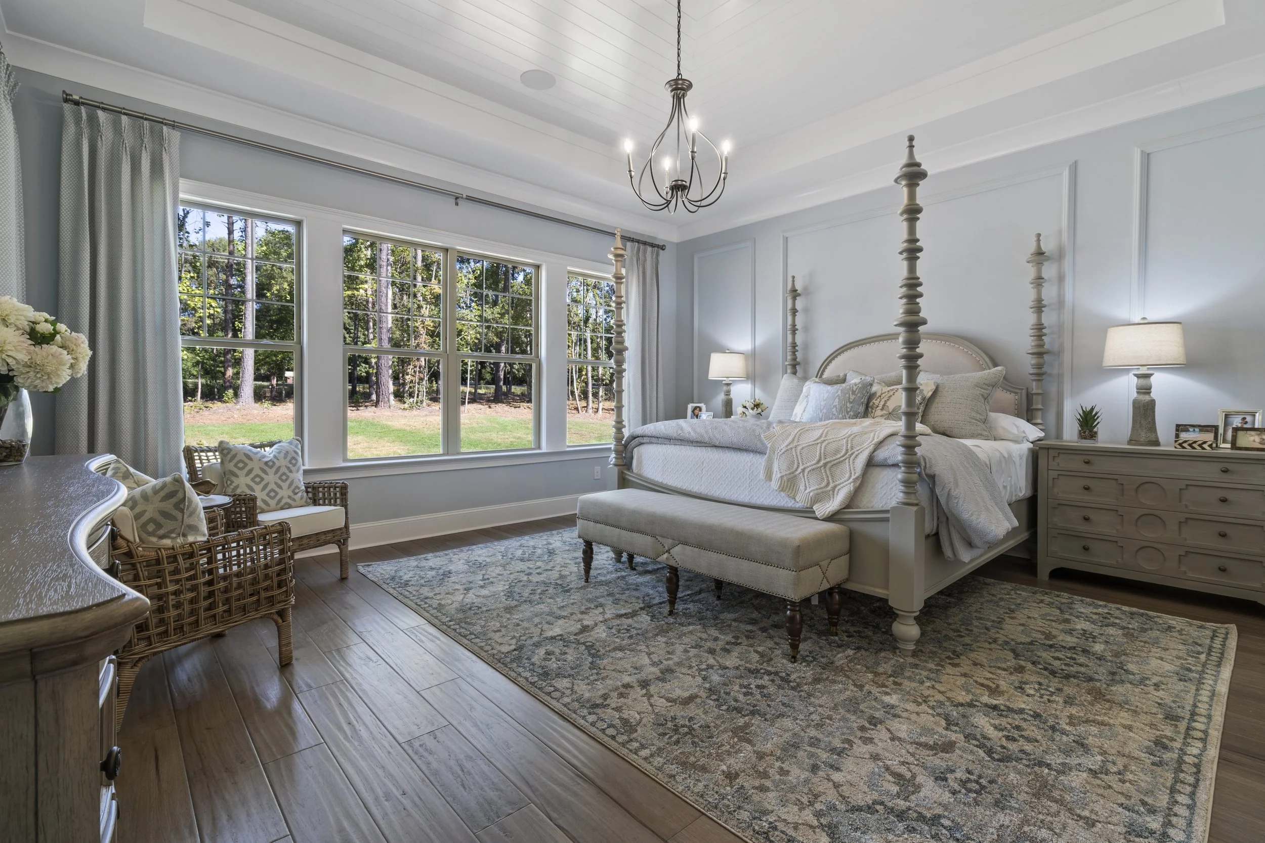 Bright bedroom with large window overlooking a wooded yard, four-poster bed with white bedding, beige upholstered bench, two nightstands with lamps, and a patterned area rug.