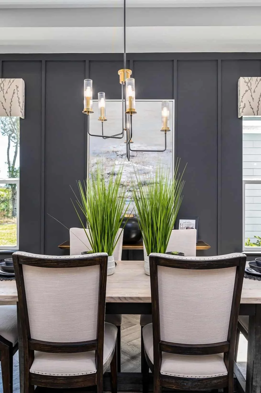 A dining room with a wooden table, four upholstered chairs, two large green plants in white planters, dark gray paneled walls, a chandelier with multiple light fixtures, windows with curtains, and a framed picture or mirror behind the plants.