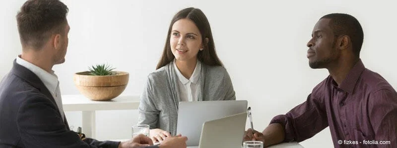 Drei Menschen im Gespräch in einem modernen Büro, eine Frau mit Laptop, zwei Männer im Gespräch.
