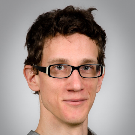 Portrait of a young man with curly brown hair, glasses, and fair skin, looking at the camera against a gray background.