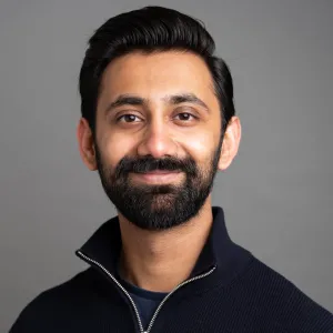 A man with dark hair and a beard, wearing a black zip-up sweater, smiling against a gray background.