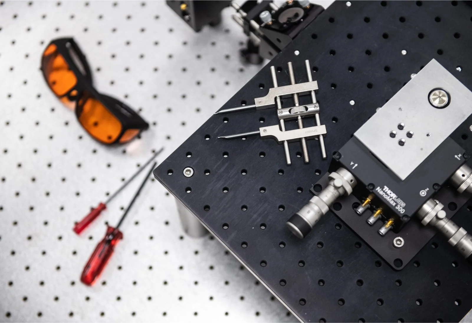 Laboratory or workshop workbench with tools including screwdrivers, a clamp, and precision equipment on a perforated black surface. Safety glasses are visible in the background.