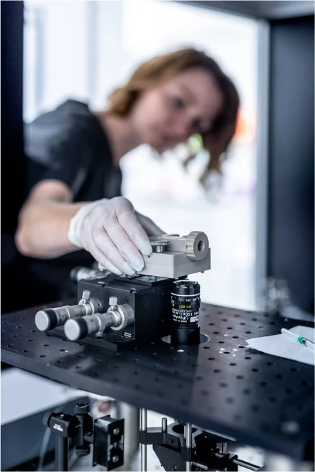 Scientist in a laboratory setting adjusting a piece of microscope equipment on a black perforated optical table, with a blurred background and window behind.