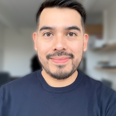 A man with short black hair, a beard, and a mustache smiling at the camera, wearing a dark blue shirt, in a modern indoor setting with blurred background.