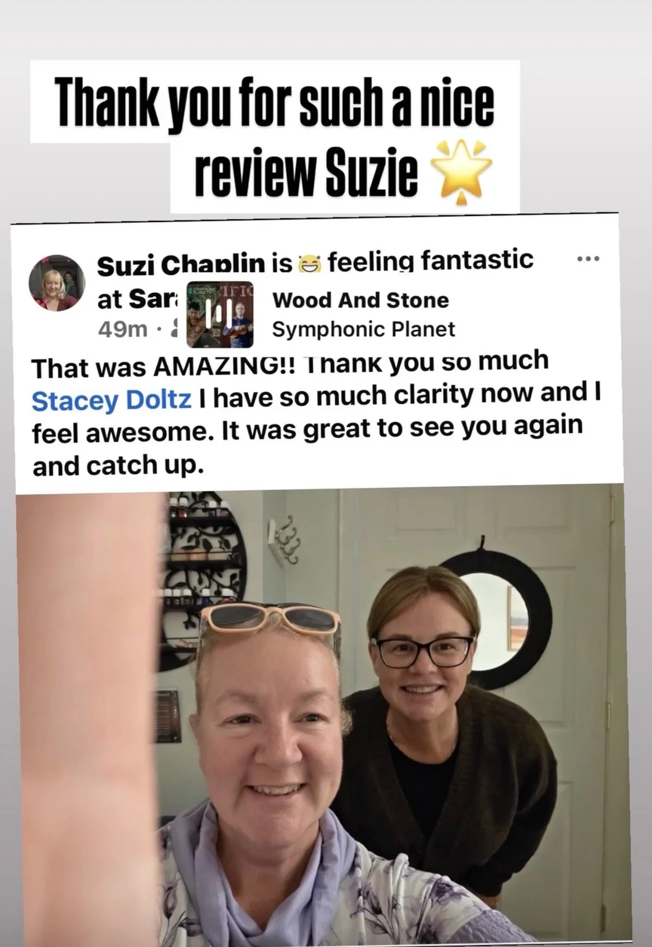 A smiling woman with glasses and pastel-colored shirt takes a selfie with a woman wearing glasses and a dark sweater, posing in front of a light-colored door with a round mirror.