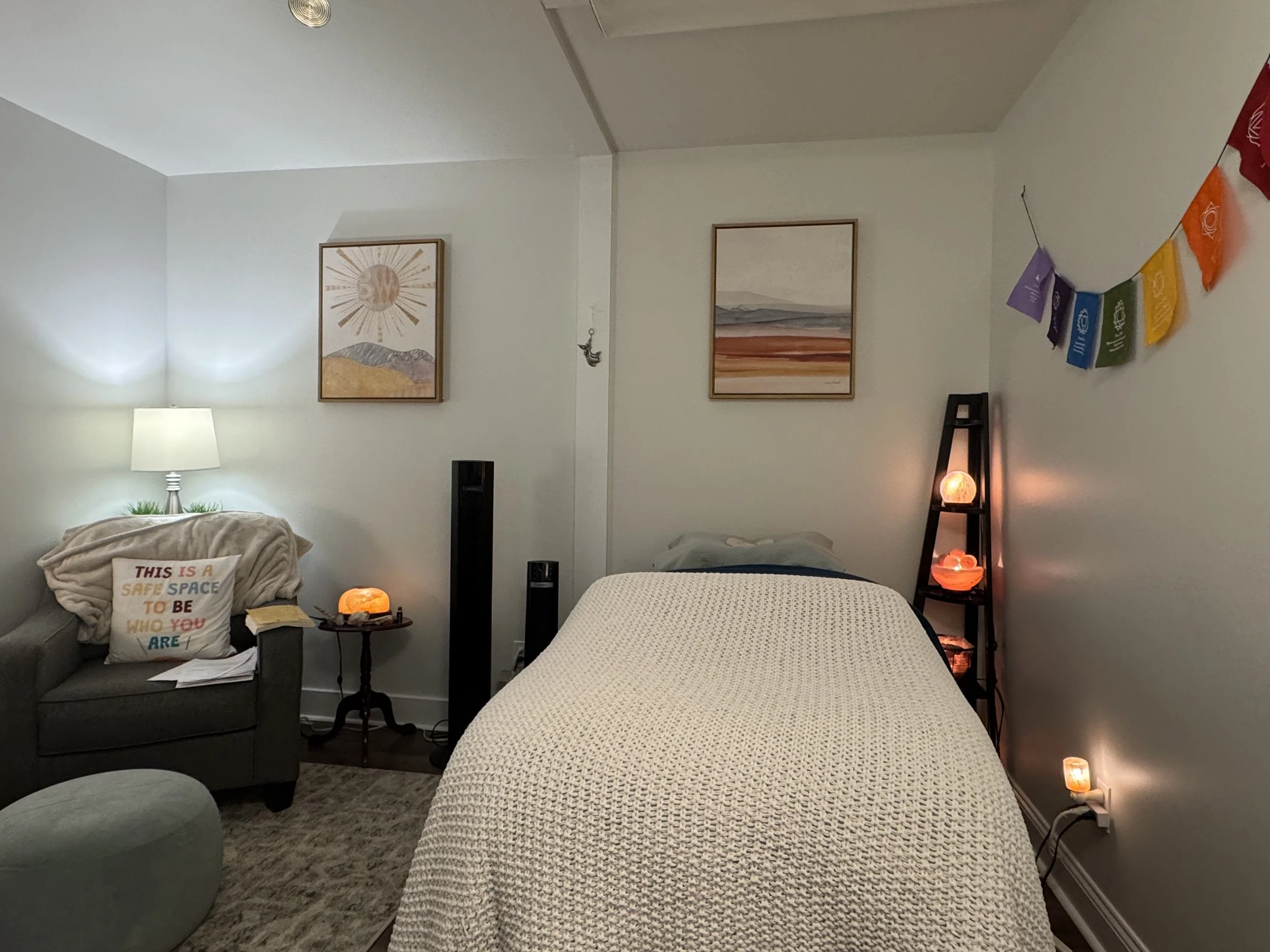 A bedroom with a bed, a gray armchair with colorful pillow, a lamp, framed artwork, and colorful prayer flags on the wall.