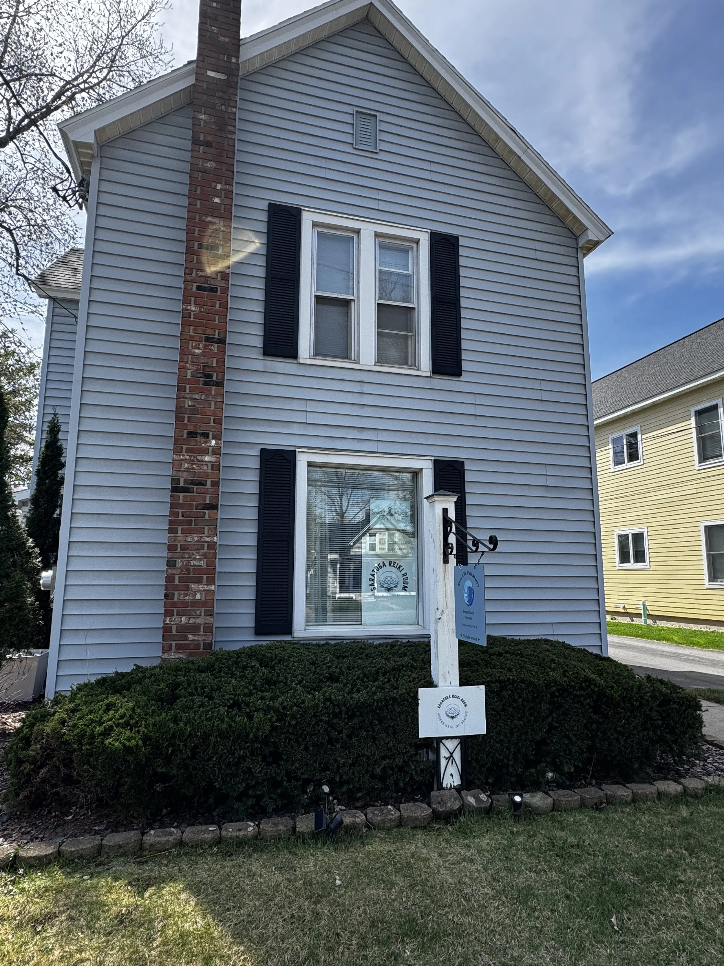 A two-story house with light blue siding, white trim, and black shutters. There is a brick chimney on the left side and a glass front door with a sign for a Reiki business. The house is surrounded by bushes and a small stone border.