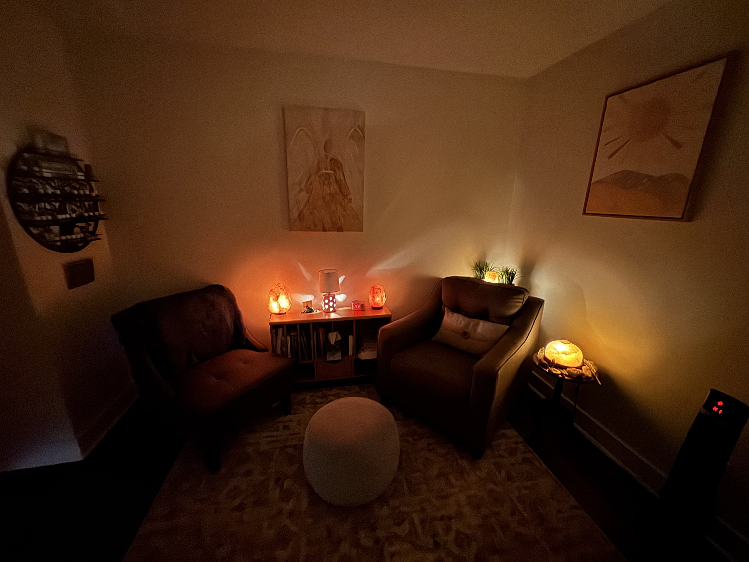 A cozy living room with dim lighting, featuring two armchairs, a small table with Himalayan salt lamps, a bookshelf, and wall art. A soft rug covers the floor, and there's a small stool or ottoman in the center of the room.
