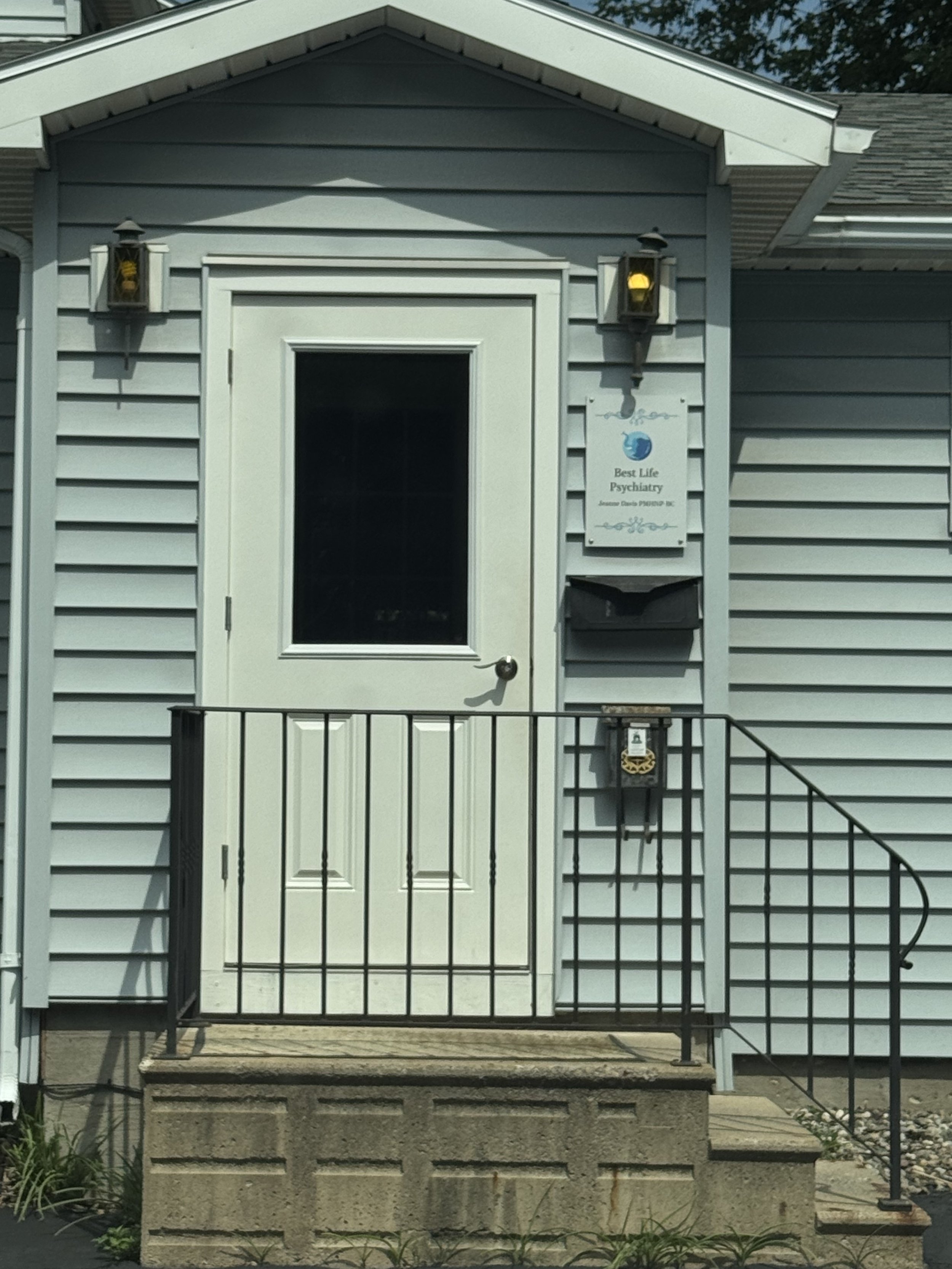 The front entrance of a building with a white door, black railing, and two wall-mounted lamps. There is a sign that reads 'Best Life Psychiatry' and a mailbox.