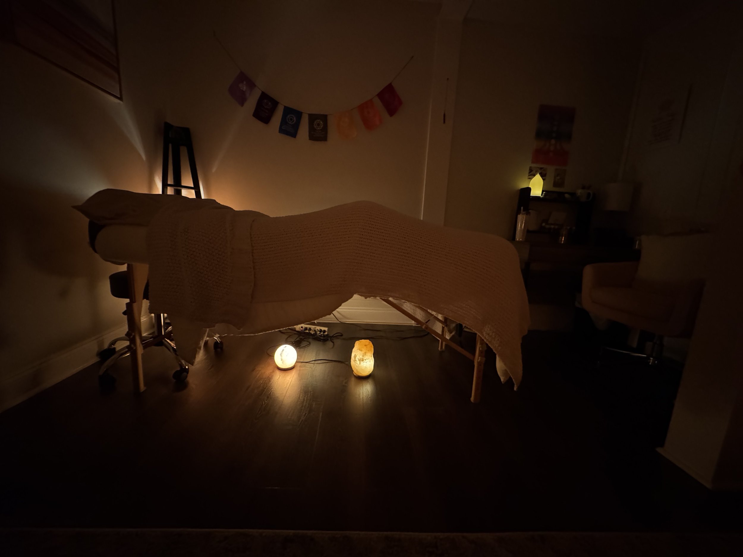 A dimly lit massage or therapy room with a massage table covered by a blanket, decorative lights on the floor, a wall with colorful flags or banners, and a small shelf with a lamp and other items. Meditation, reiki, angels, spirit, healing, wellness,