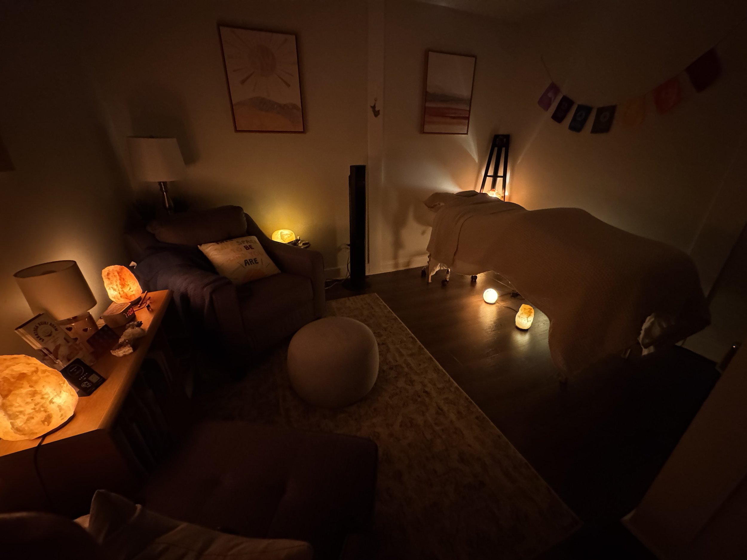 Dimly lit living room with salt lamp lights, a salt lamp on the side table, salt lamps on the floor, a salt lamp on the desk, a sofa with a cushion, a small round ottoman, and paintings on the wall. chakras, energy field,medium, psychic, angel card