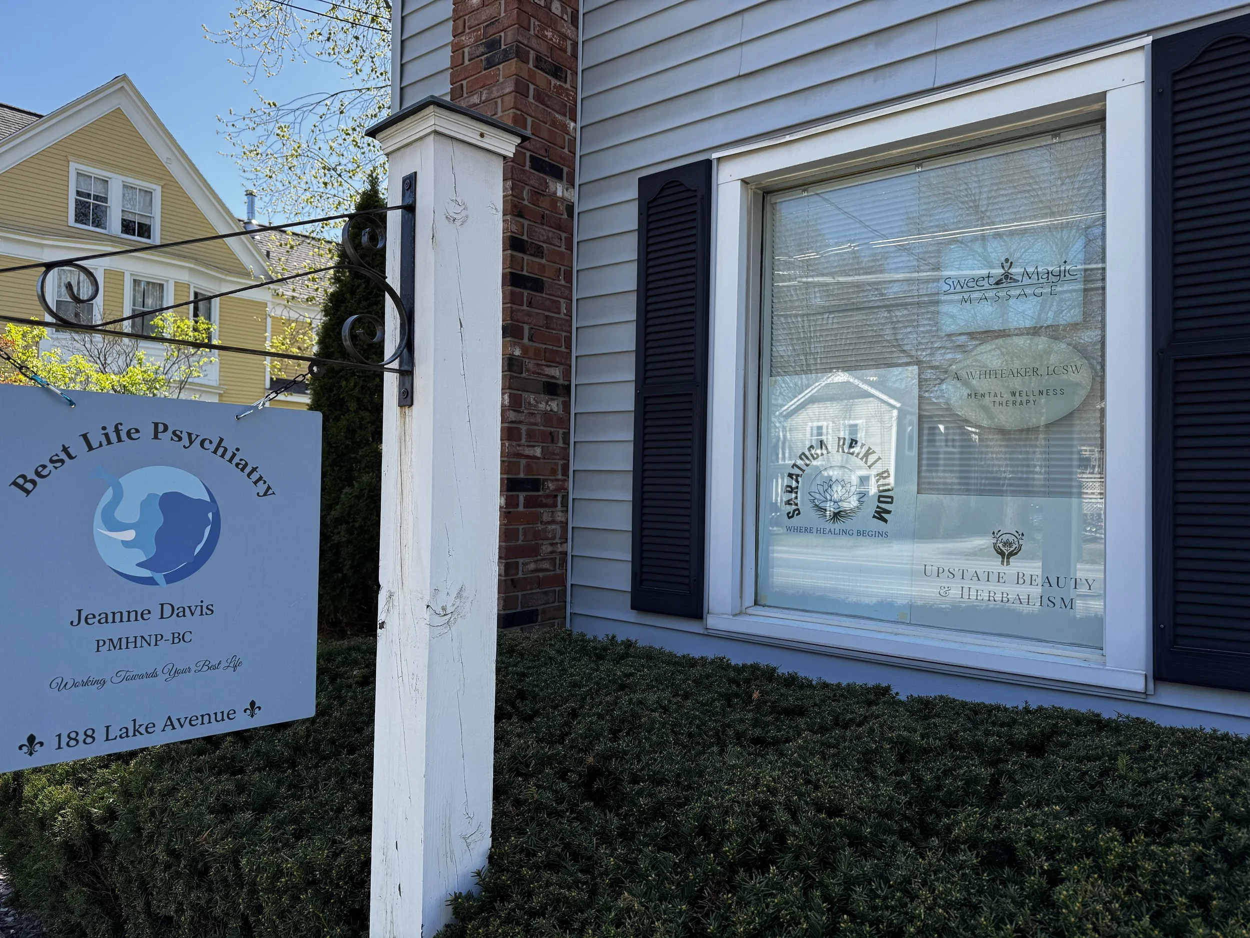 A storefront window with signs for wellness and psychotherapy services. A white wooden post and a sign with blue and black text are visible, along with shrubs and a partial view of a yellow house in the background.