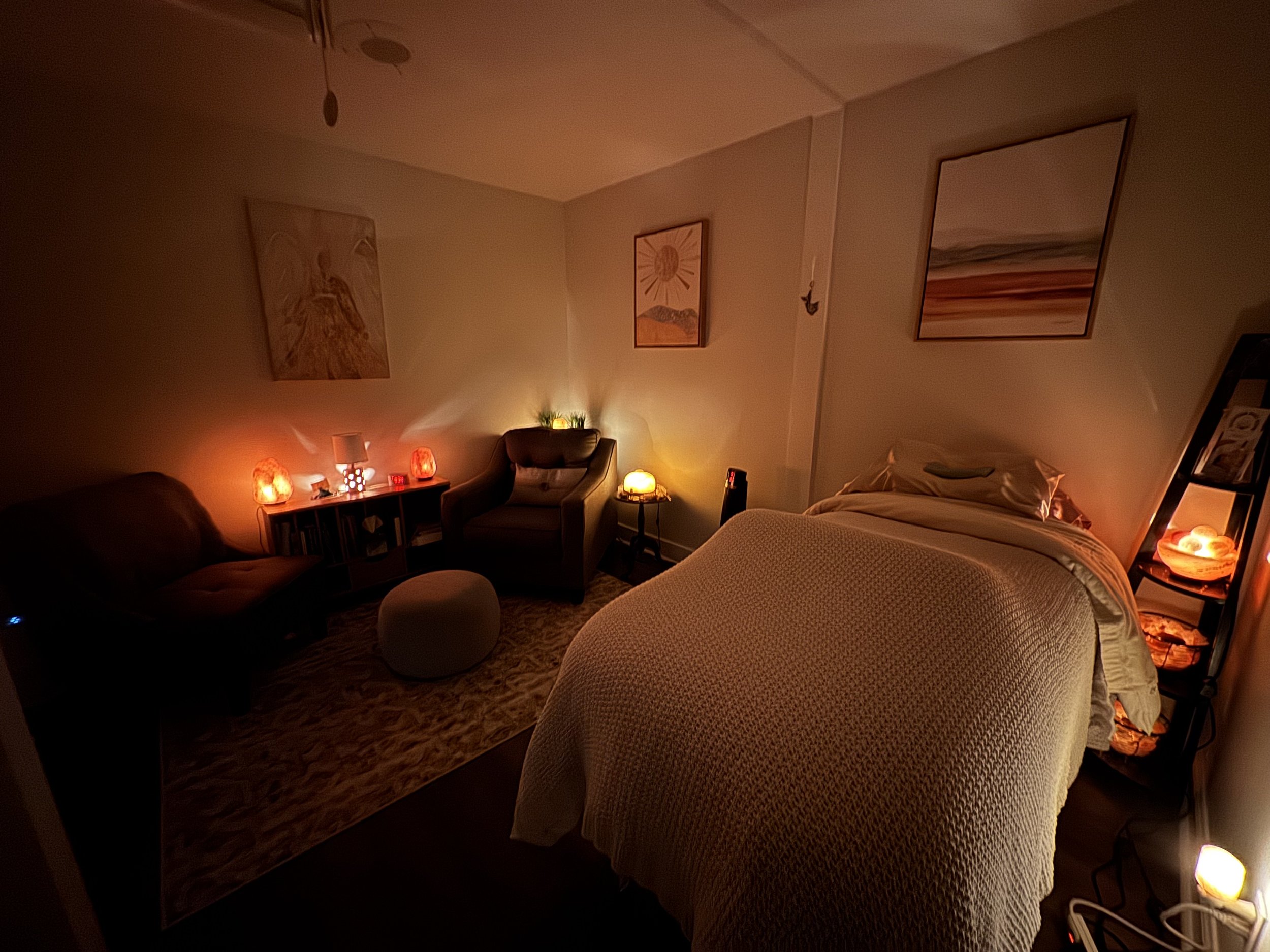 Cozy therapy room with dim lighting, massage table, armchair, and decorative lamps.