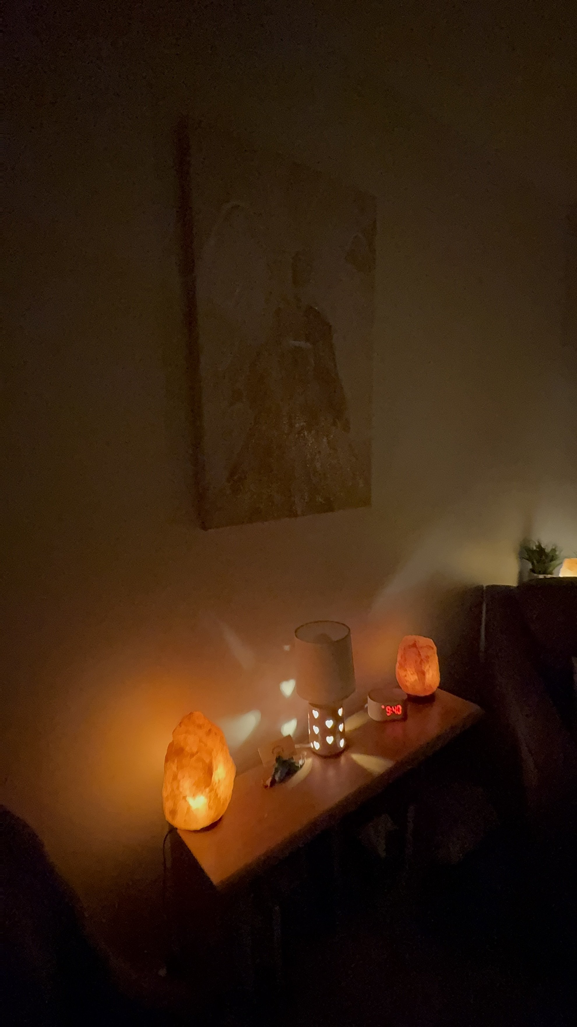 Dimly lit living room with salt lamps, a lamp with heart-shaped cutouts, a digital clock, and a Himalayan salt lamp on a wooden table. A small painting hangs on the wall.