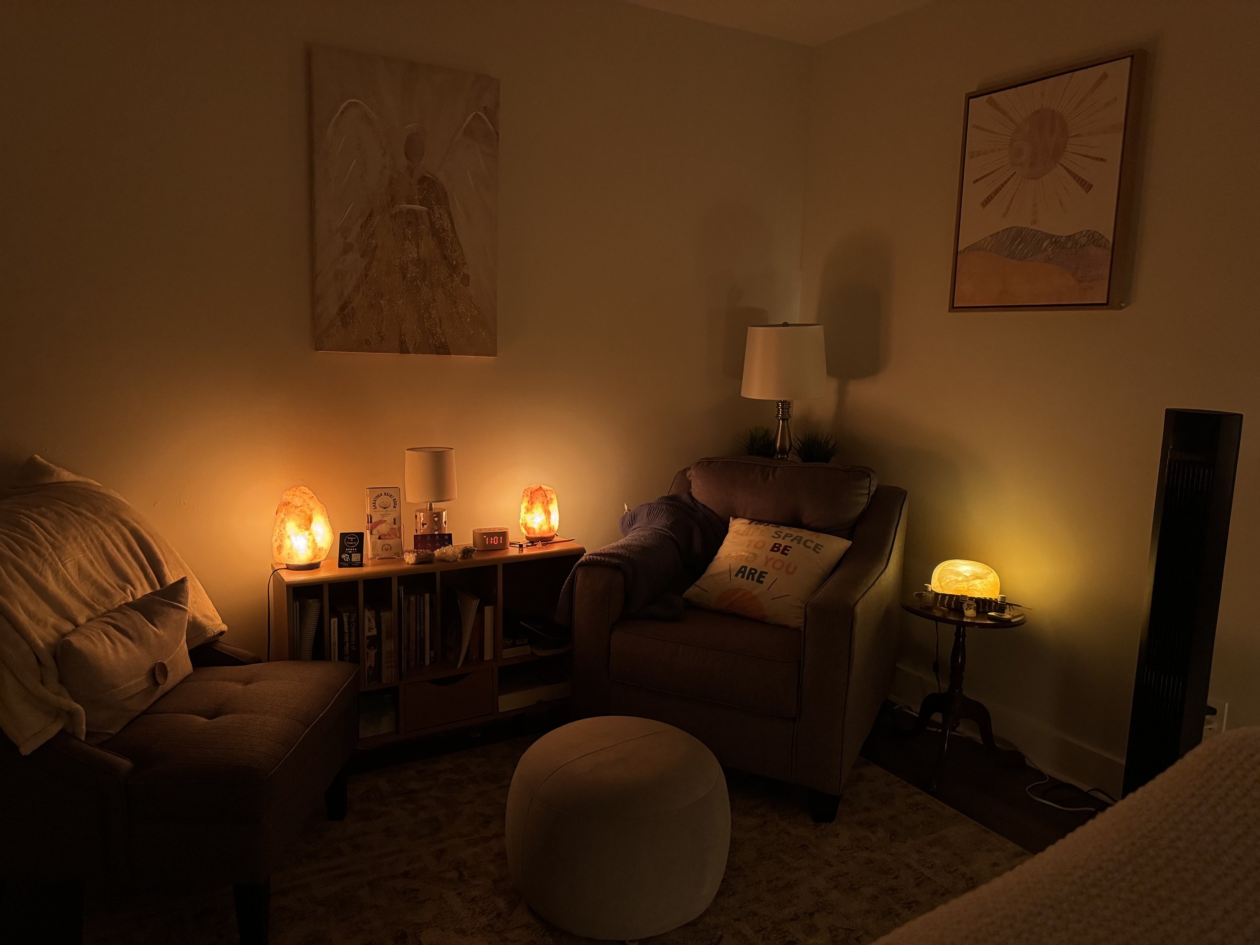 Living room with dim lighting, salt lamps, a sofa, armchair, bookcase, and framed artwork on the wall.