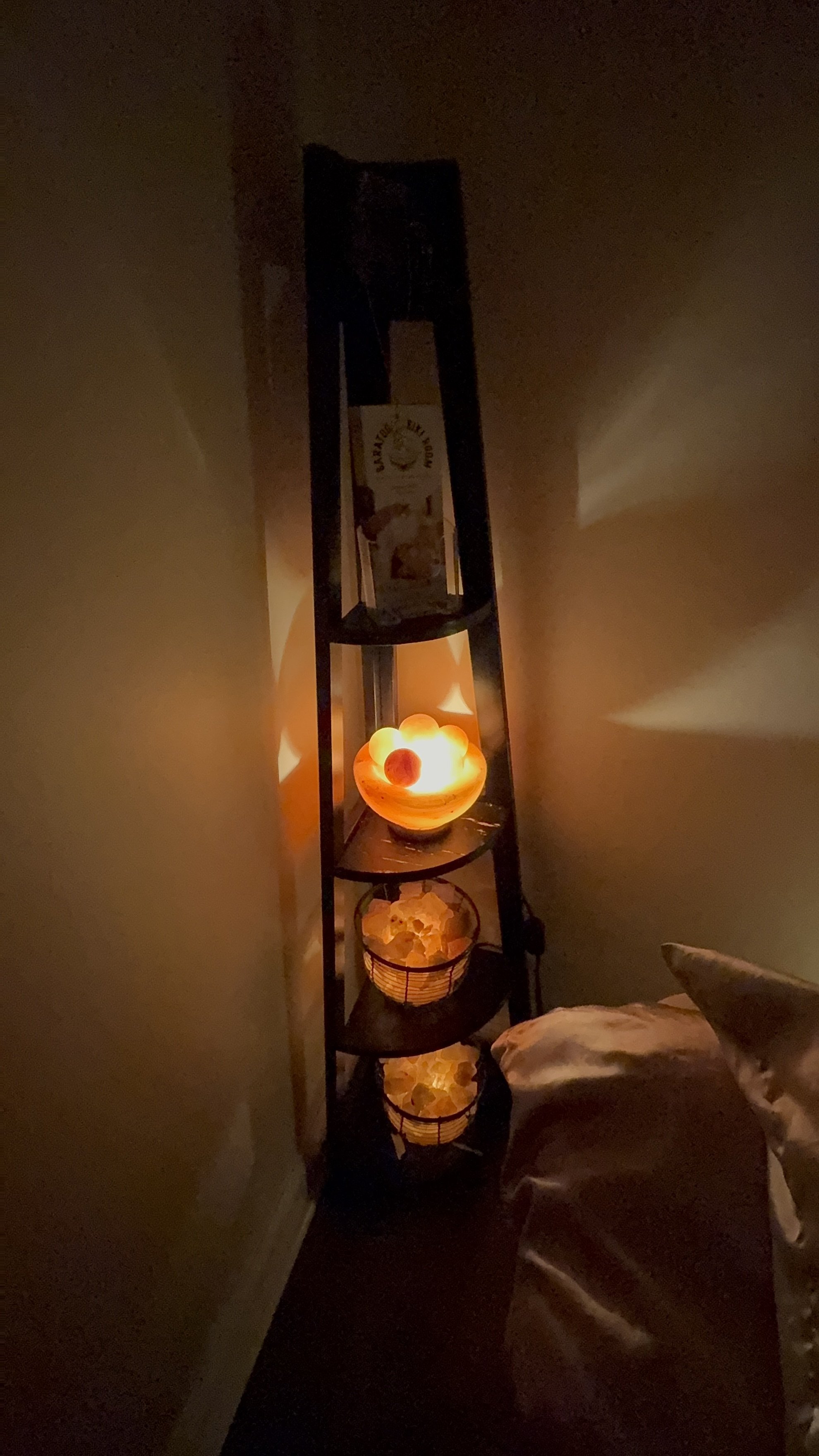 A corner of a room with a black metal corner shelf illuminated by warm orange light from Himalayan salt lamps. The top shelf has a box of Himalayan pink salt and the middle shelf has a bowl with glowing orbs. The bottom shelf holds two salt lamps.