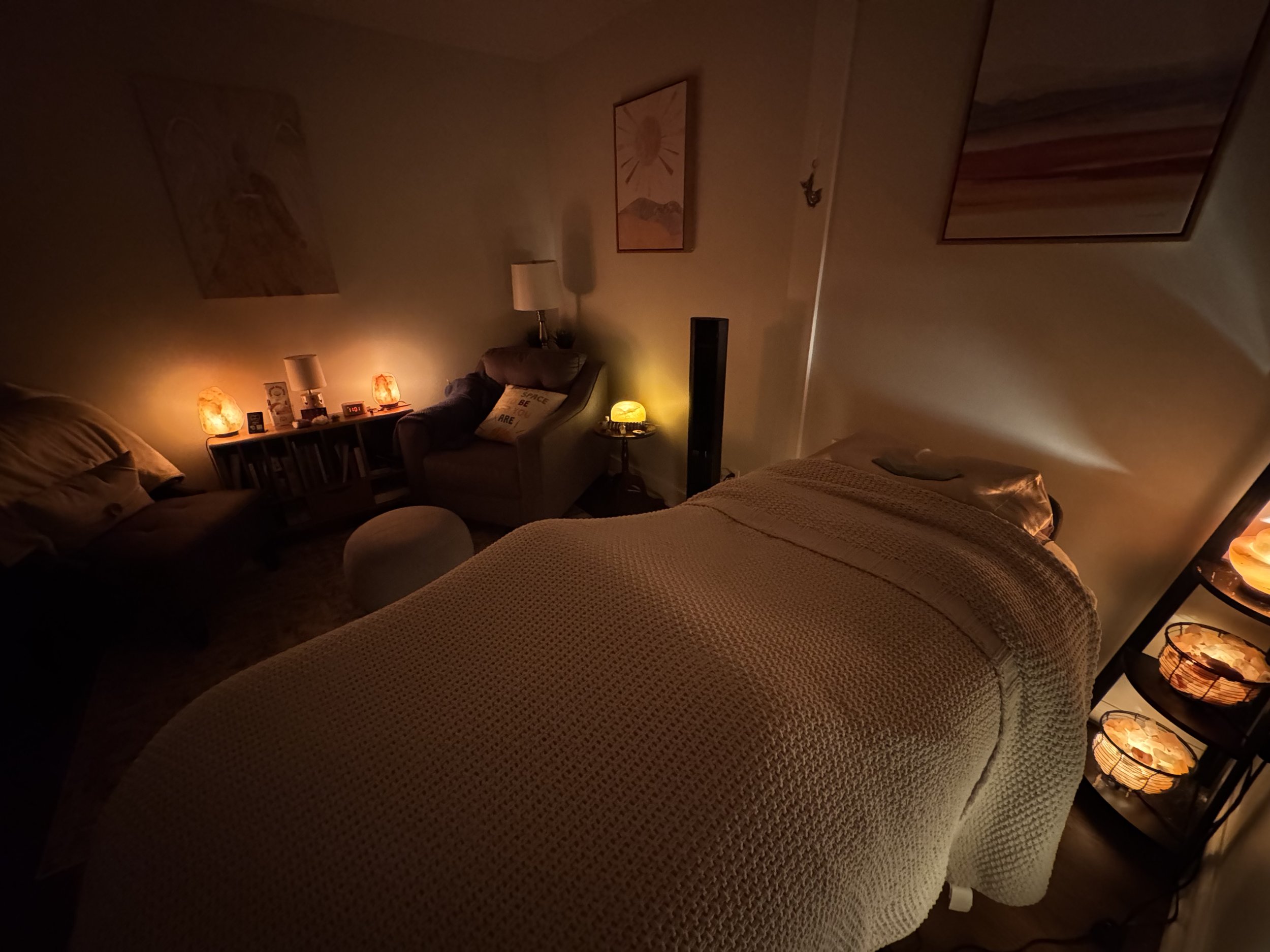 A dimly lit bedroom with a bed and a massage table, illuminated by warm, soft lamps and candles.