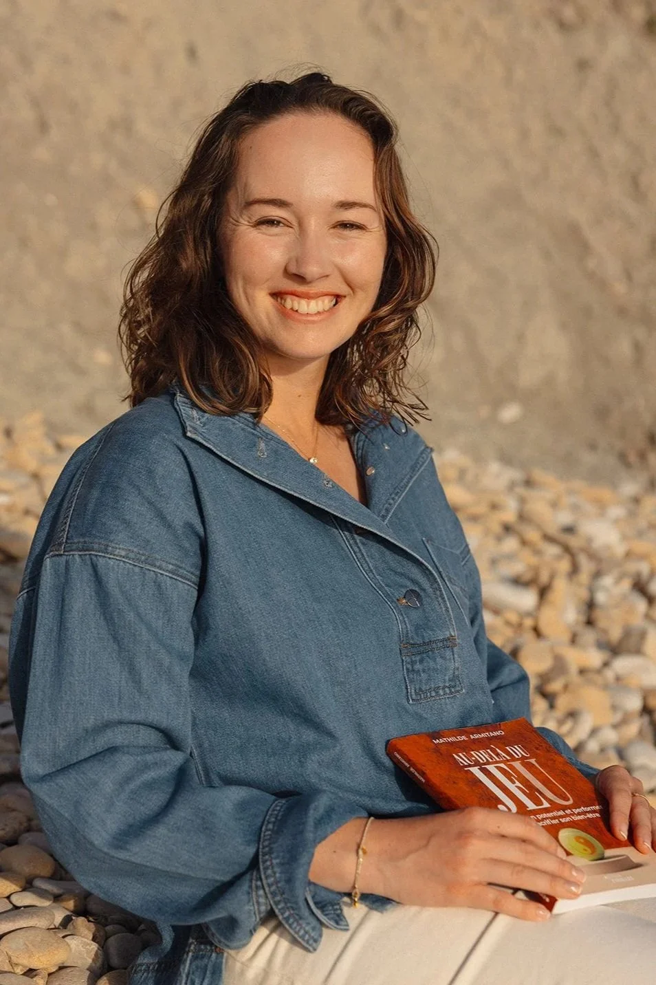 Une femme souriante avec des cheveux bruns bouclés, portant une veste en denim, assise sur des cailloux, tenant un livre rouge intitulé 'Au-delà du feu'.