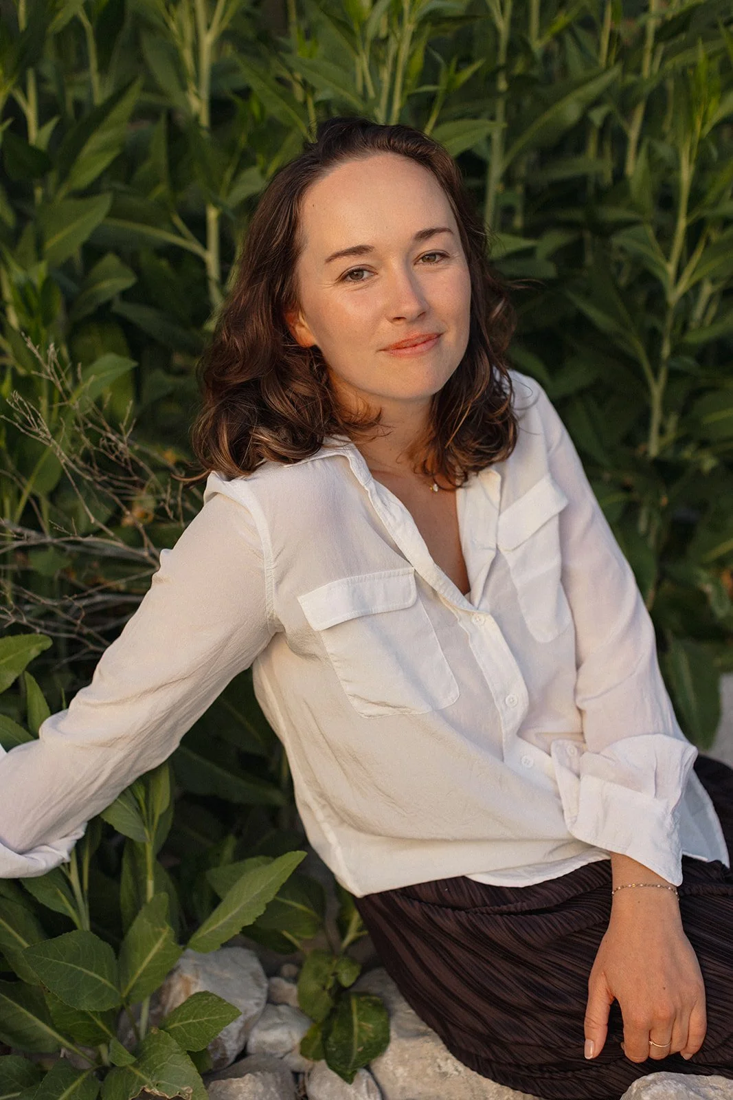 Jeune femme assise près de plantes vertes, portant une chemise blanche et un pantalon foncé, avec une expression calme et un léger sourire.