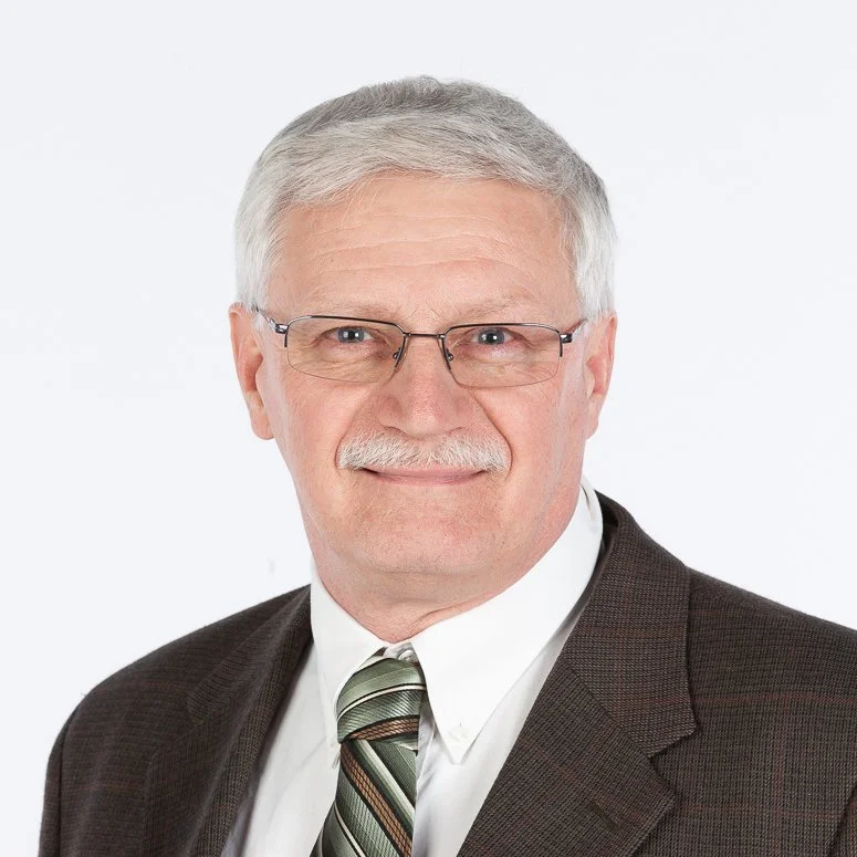 A professional portrait of an older man with glasses, gray hair, and a mustache, wearing a dark brown suit jacket, white shirt, and striped tie, smiling against a plain white background.