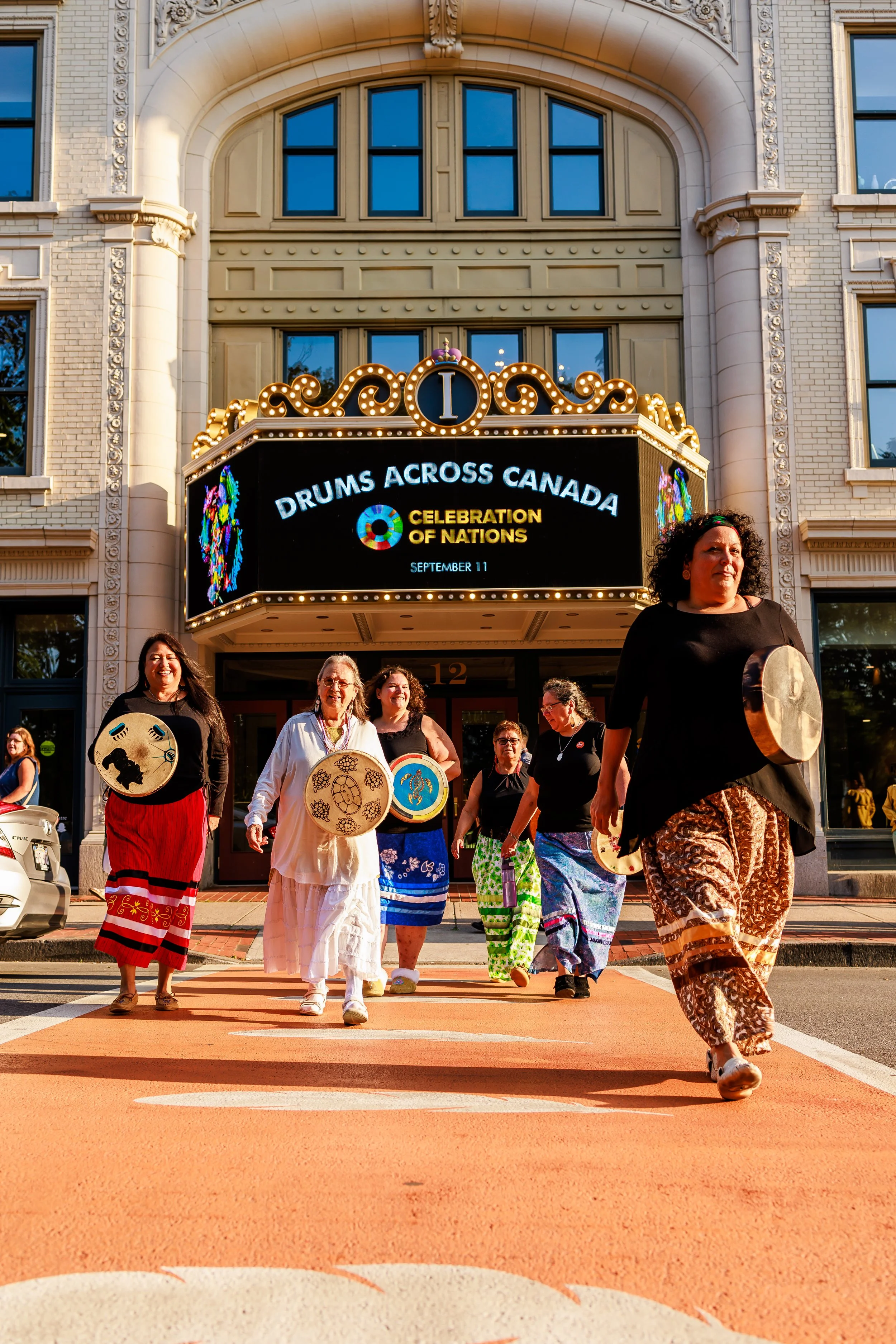 Group of women in colorful skirts and black tops, carrying drums, walking across a crosswalk in front of a building with a marquee reading 'Drums Across Canada' during a celebration event.