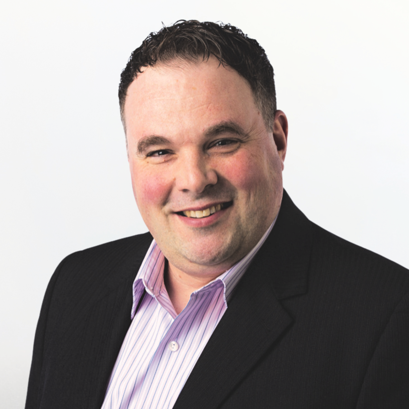 A professional headshot of a smiling man with short dark hair, wearing a black suit jacket and a pink striped dress shirt, against a plain white background.