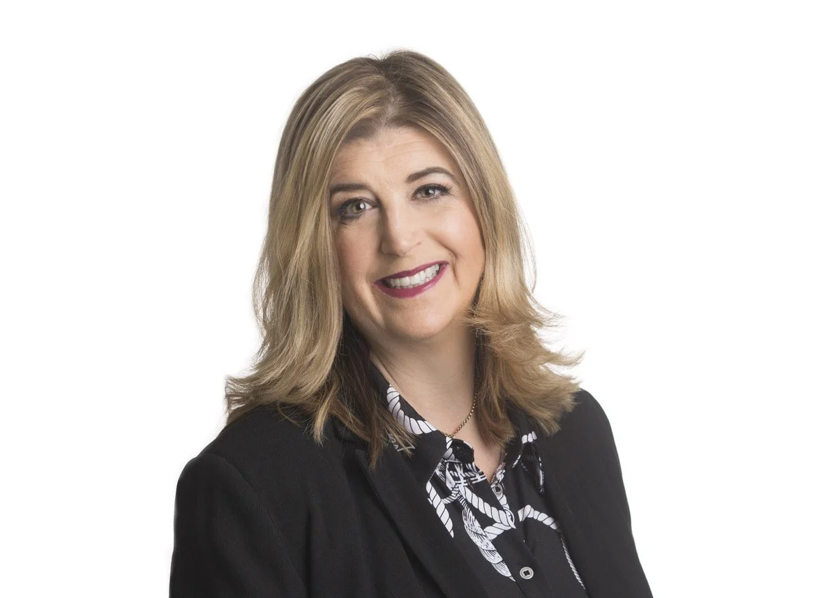 A woman with shoulder-length blonde hair, smiling, wearing a black blazer over a patterned blouse, against a white background.